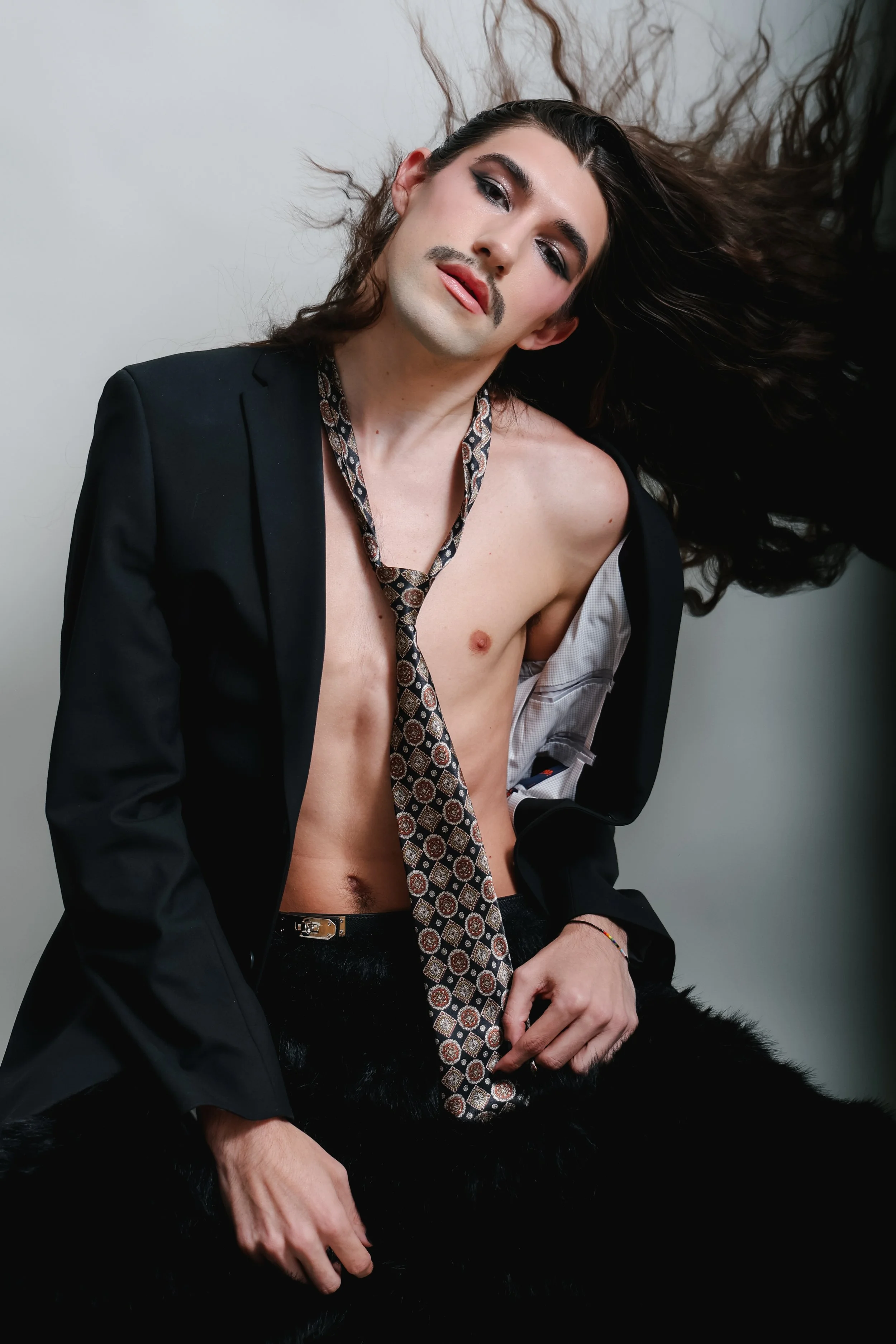 A person with long dark hair, makeup, and a mustache, wearing a black blazer, a patterned tie, and a partially open shirt, sitting against a plain light gray background with a black furry item on their lap.