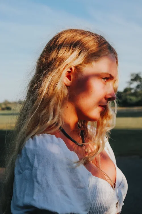 Side profile of a young woman with long blonde hair, wearing a white top and a black necklace, outdoors during sunset.
