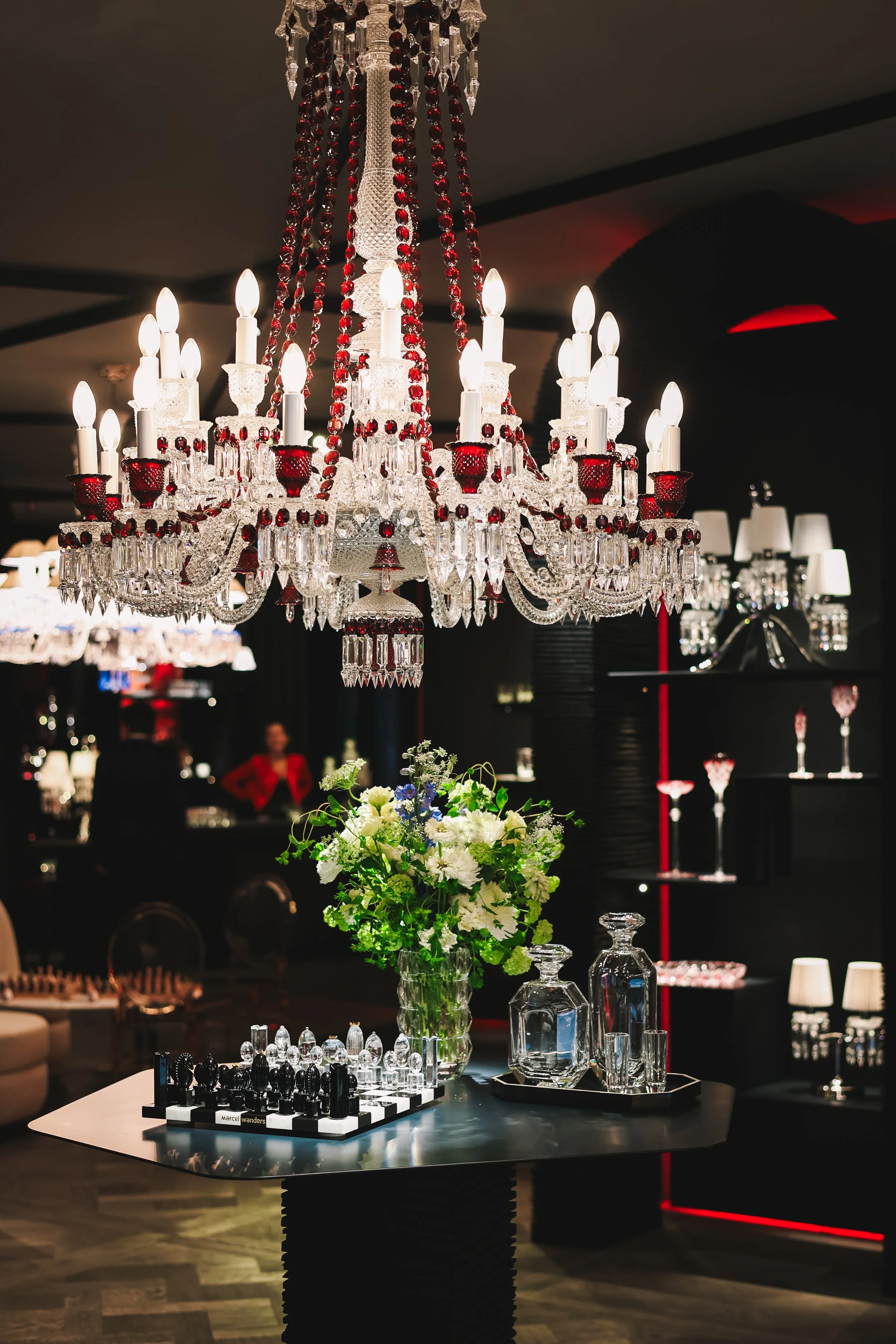 Elegant chandelier with red and clear crystals hanging above a table with a floral centerpiece, a chess set, and glass decanters in a luxurious setting.