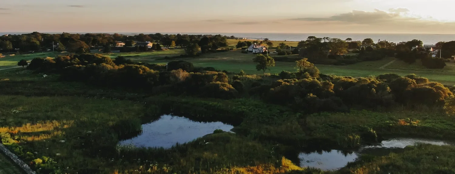 Sunset over a rural landscape with fields, trees, houses, a pond in the foreground, and the ocean horizon in the distance.