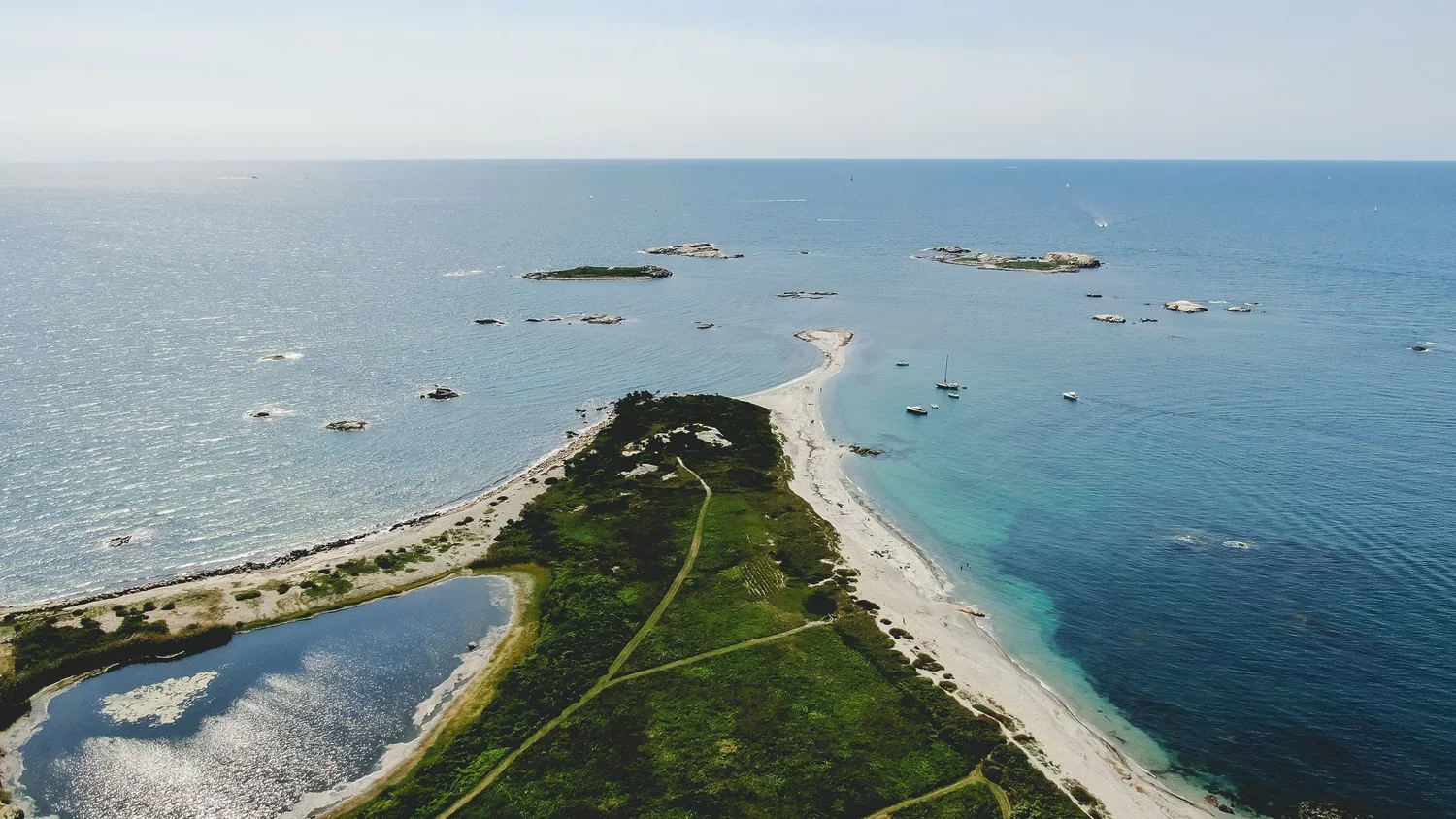 Aerial view of a coastal area with a narrow strip of land extending into the ocean, dotted with boats and surrounded by water on both sides, with some small islands in the distance.