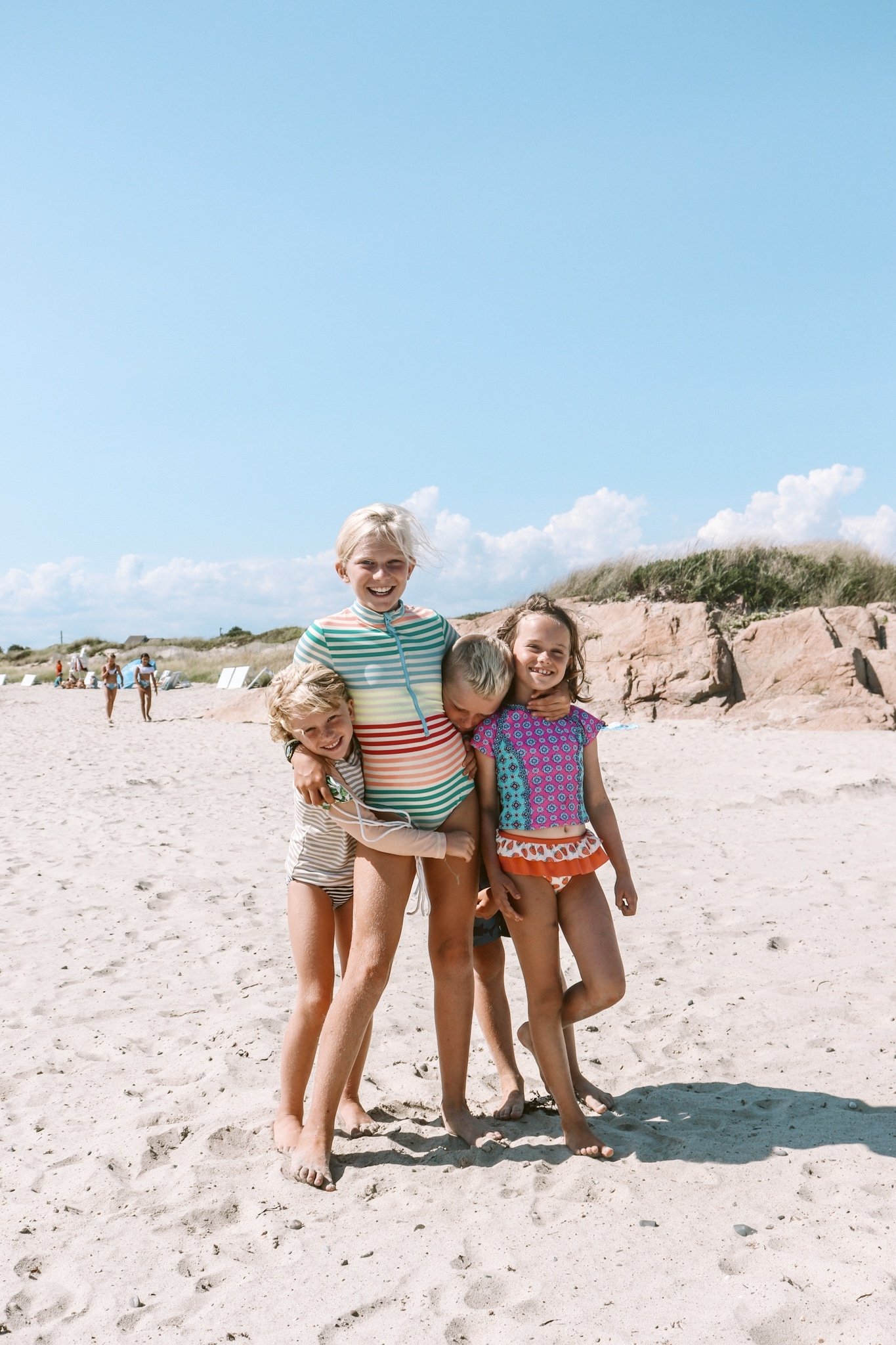 Children hugging and smiling on a sandy beach with rocks and grassy dunes in the background under a blue sky.