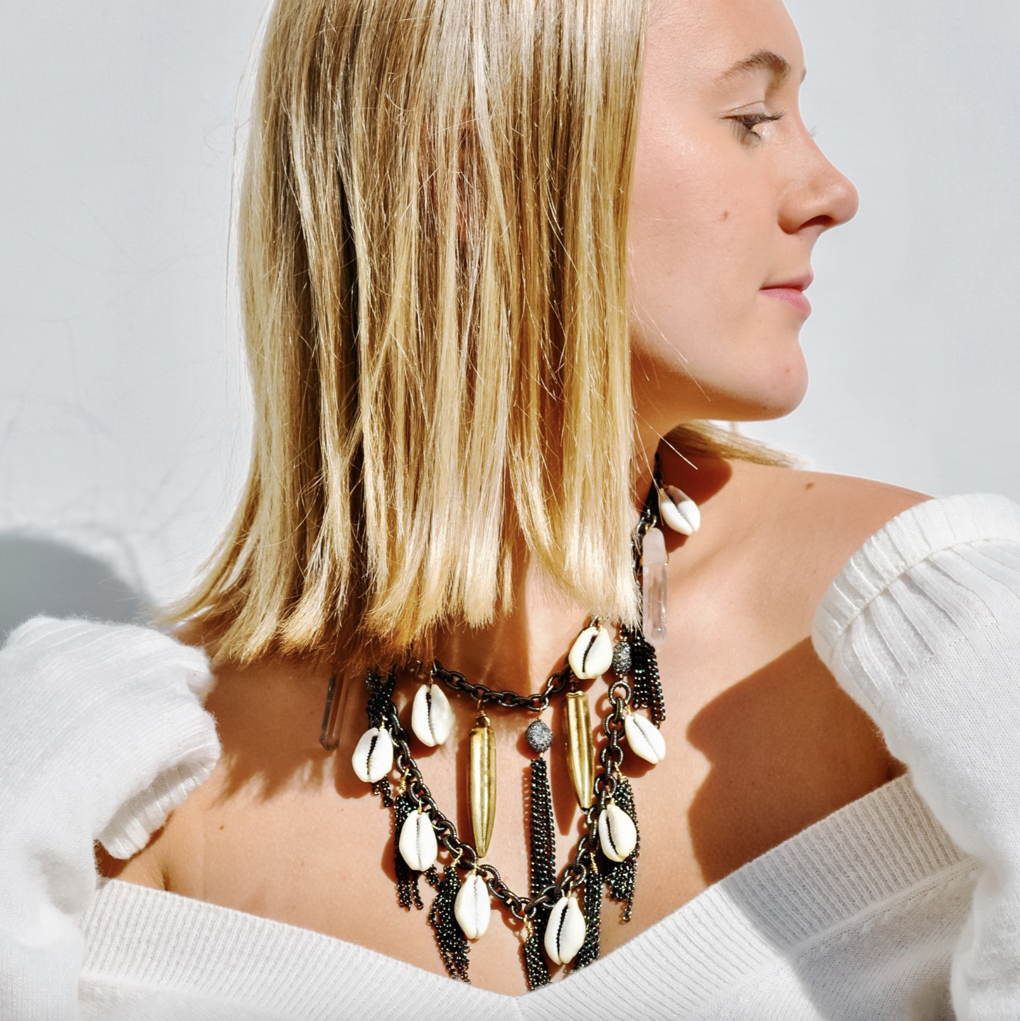 Profile of a woman with blonde hair wearing multiple layered necklaces with shells and beads, and a white off-shoulder top.