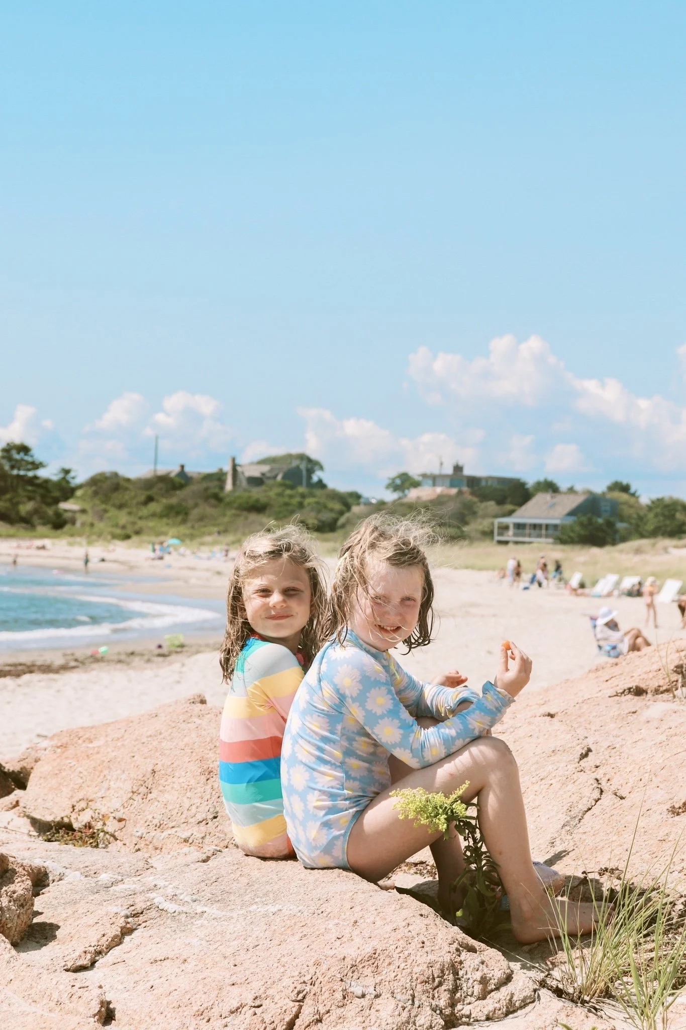 Two young girls sitting on a rock at the beach, smiling and enjoying the sunny day with blue sky and people in the background.