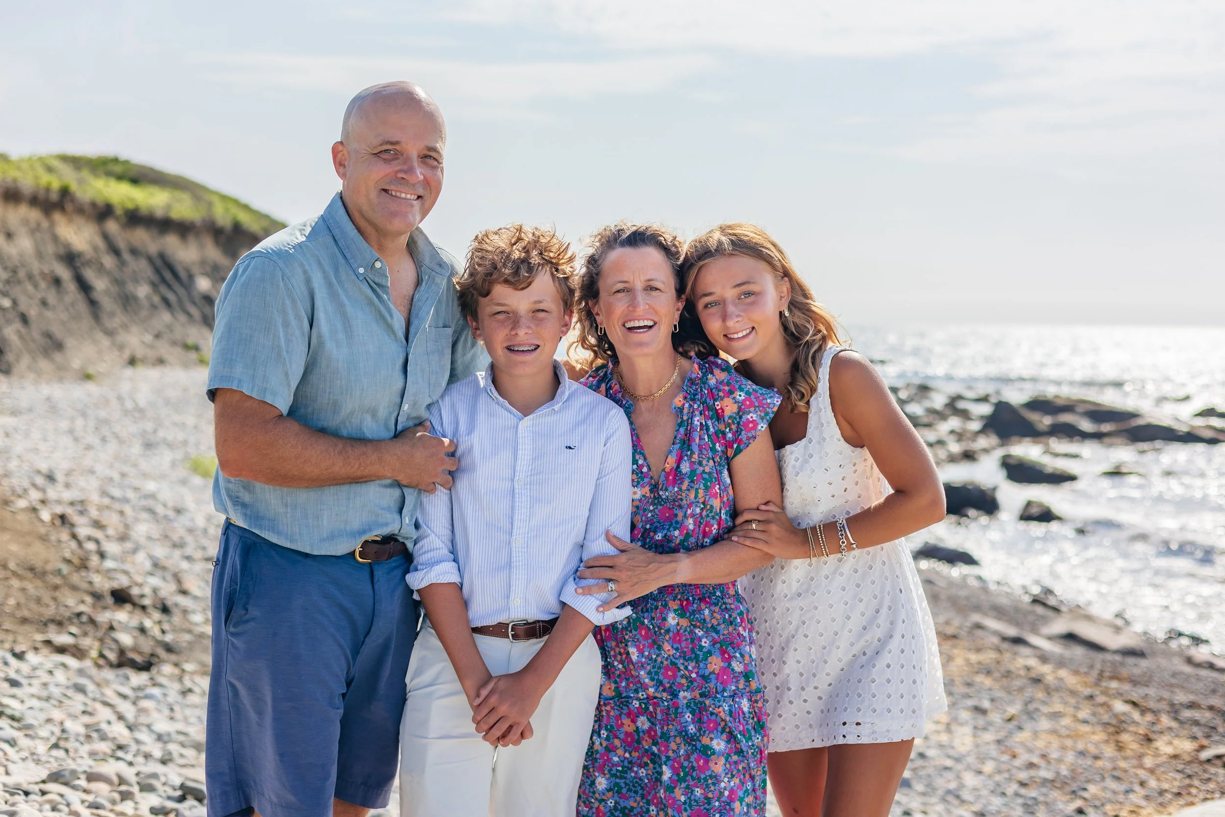 A family of five smiling and standing together on a rocky beach, with cliffs and the ocean in the background during daytime.