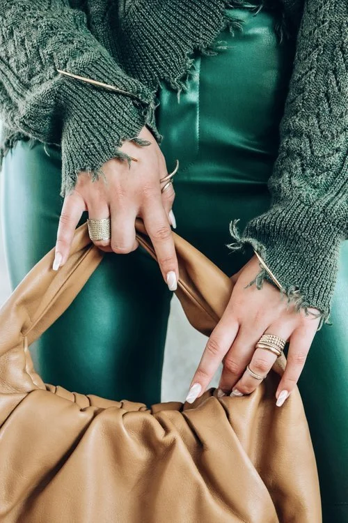 Close-up of hands holding a tan leather bag, wearing multiple rings and a green or teal leather outfit, with a gray sweater.