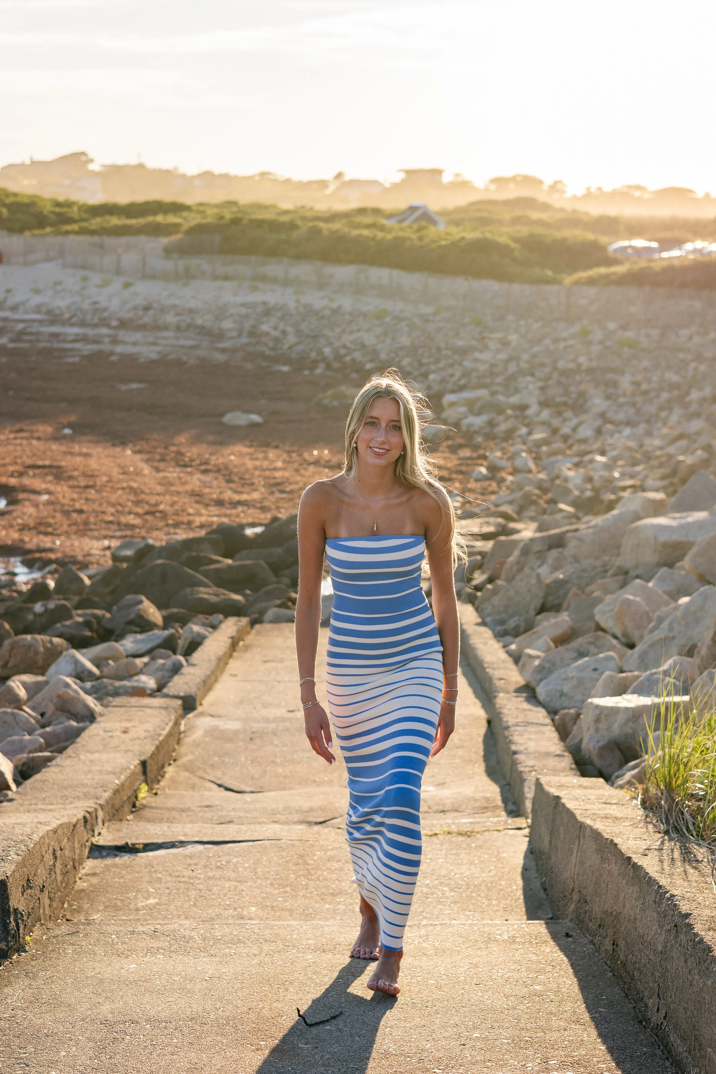 A woman in a blue and white striped dress walking barefoot on a stone path near the beach, with rocks, green hills, and parked cars in the background during sunset.
