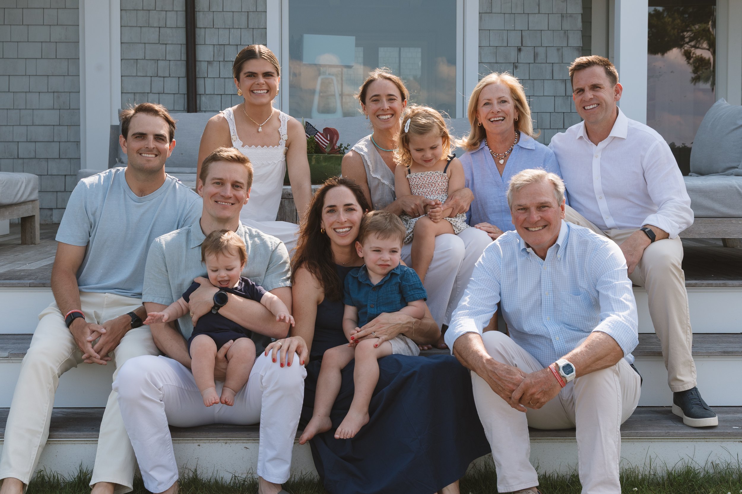 A large multigenerational family group sitting outdoors on steps in front of a house, smiling for the camera. The group includes children, adults, and seniors, dressed in casual clothing.