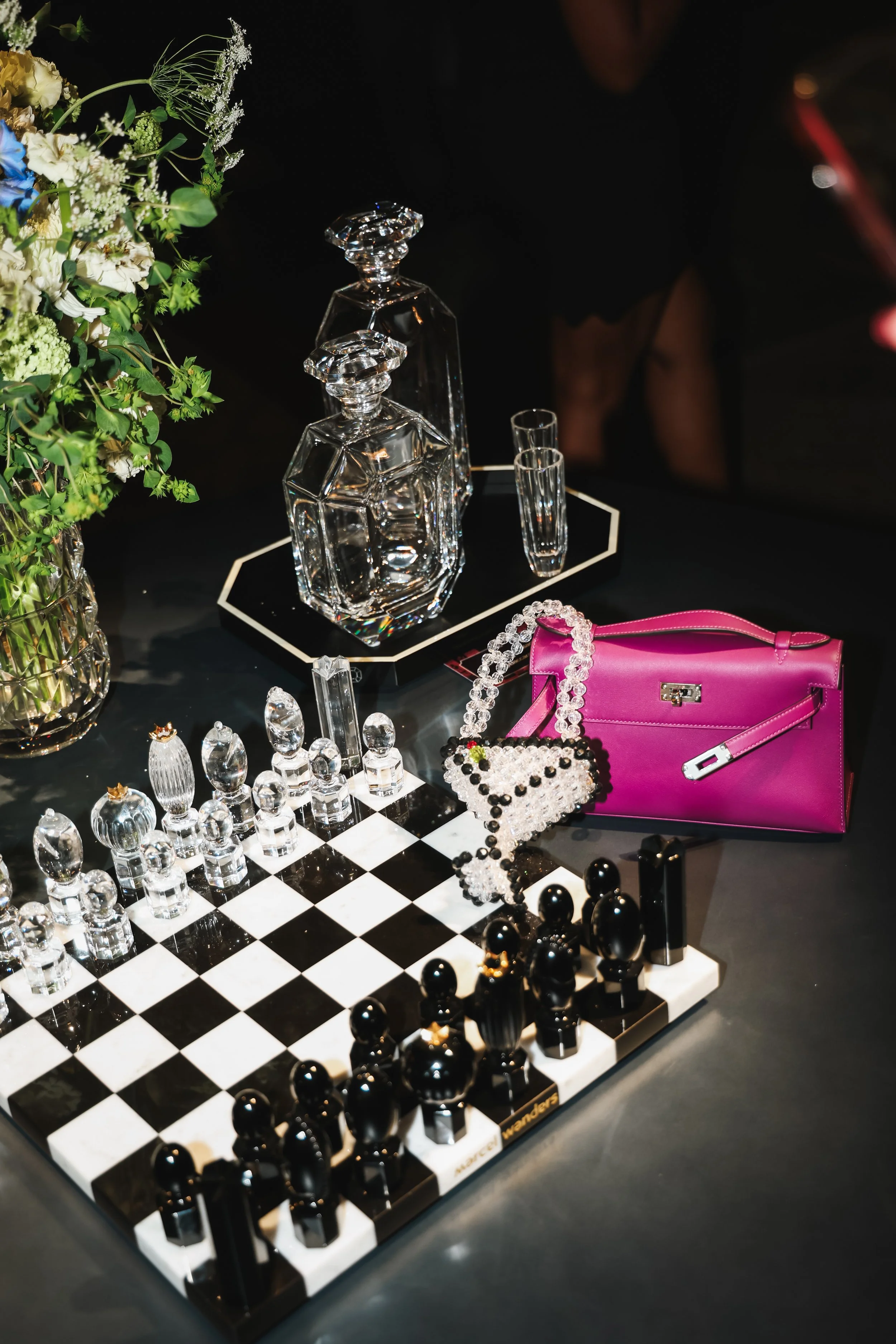 Close-up of a chessboard with glass and black chess pieces, a vase with flowers, and various jewelry including rings, earrings, and a pink purse, in a dark setting.
