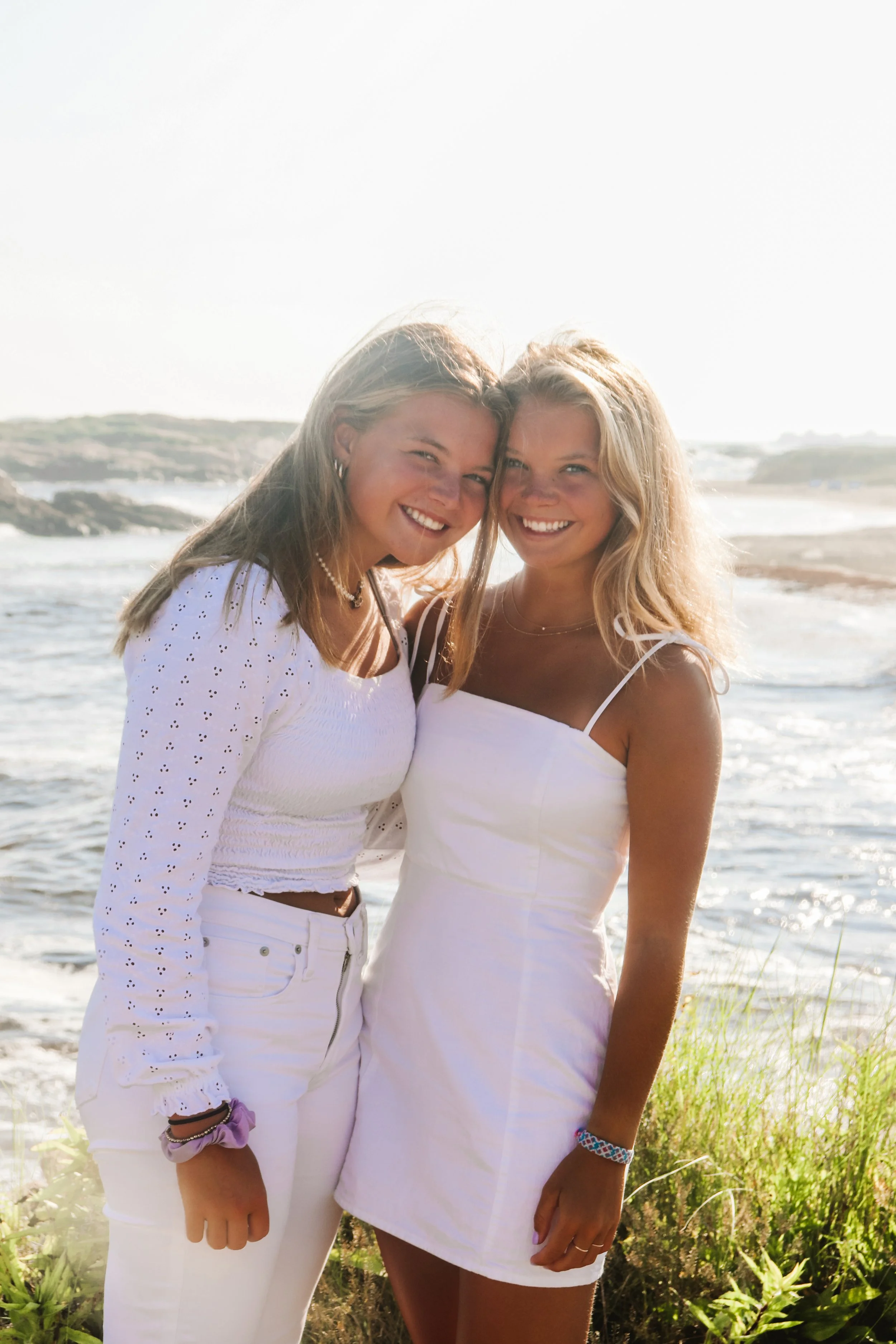 Two young women smiling at the camera on a beach, with ocean waves and rocky shoreline in the background, both dressed in white summer clothing.