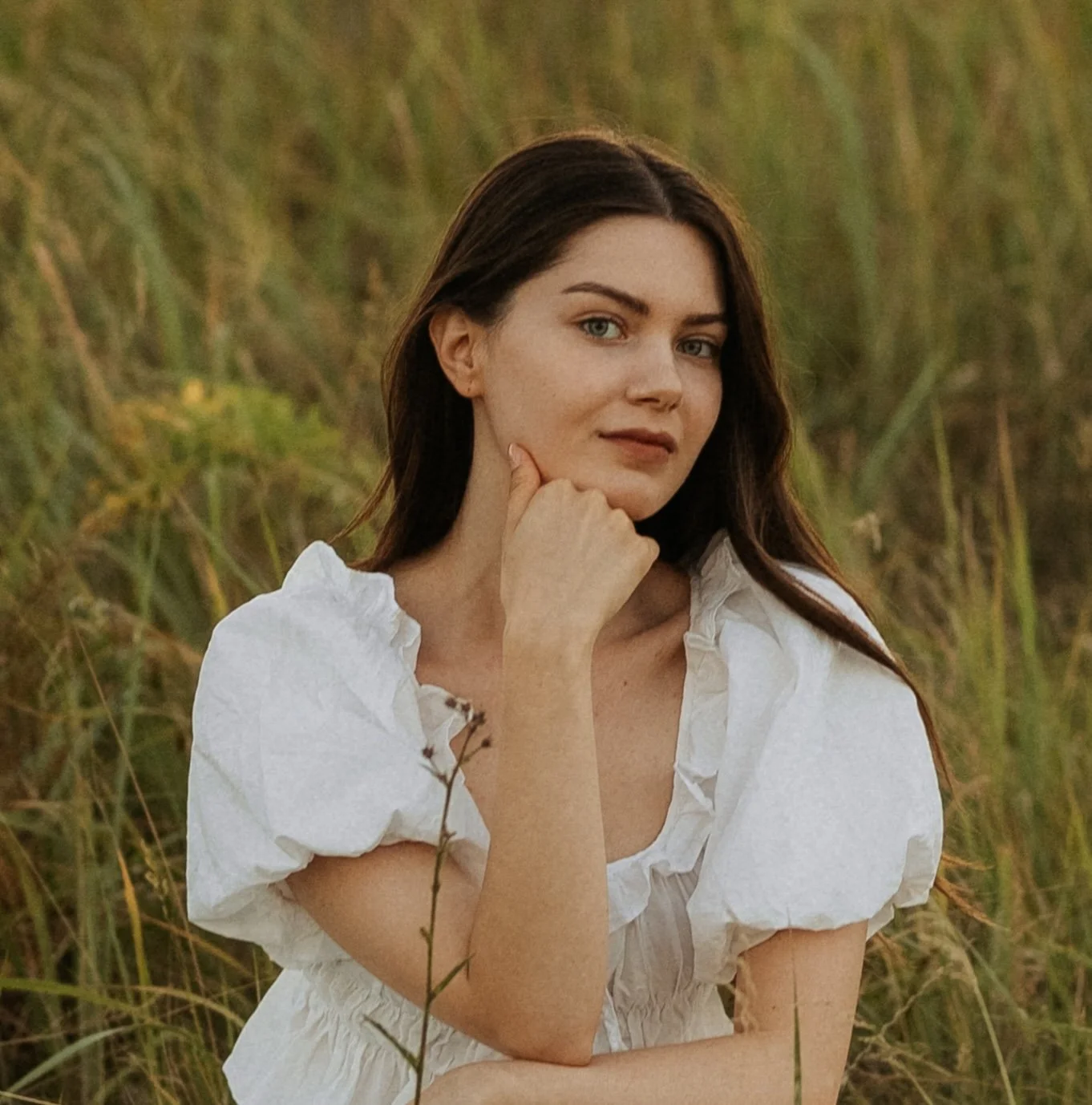 A young woman with dark brown hair in a white blouse, standing in a grassy field, looking at the camera with her hand resting under her chin.