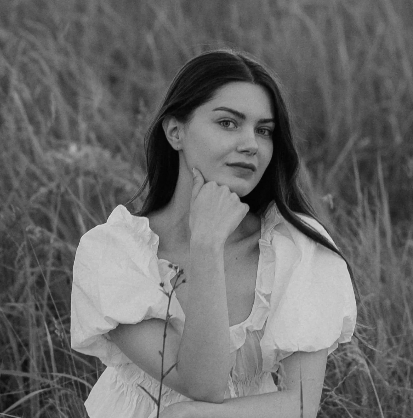 A young woman with dark brown hair in a white blouse, standing in a grassy field, looking at the camera with her hand resting under her chin.