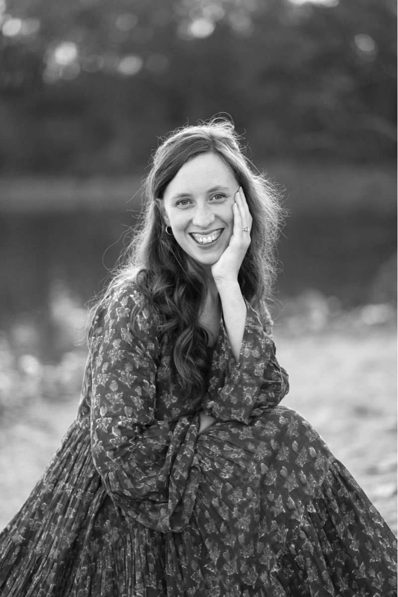 A young woman with long, wavy brown hair wearing a navy blue dress with puffed sleeves, smiling outdoors with greenery and sunlight in the background.