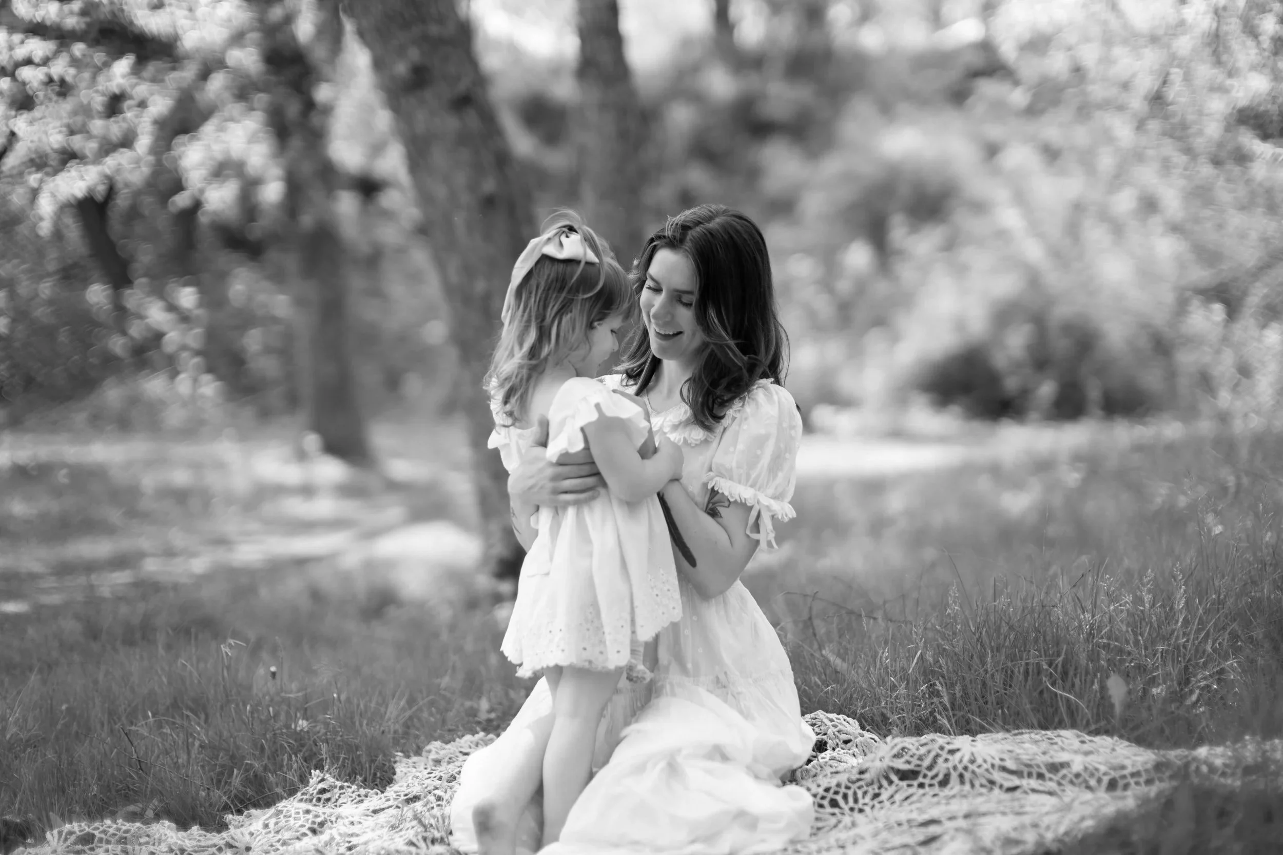A woman and a young girl sharing a joyful moment outdoors in a park or garden, sitting on a blanket surrounded by green grass and trees.