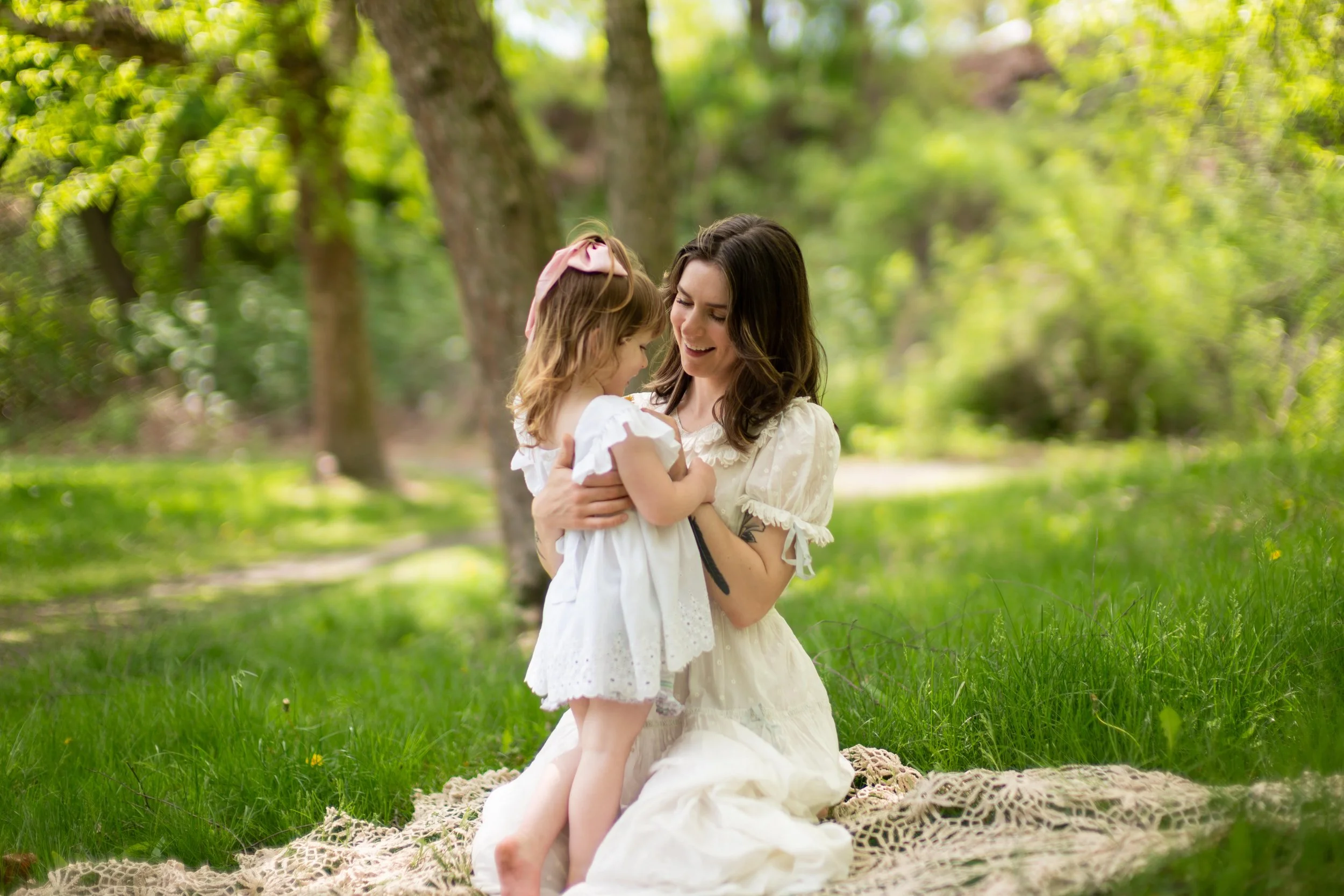 A woman and a young girl sharing a joyful moment outdoors in a park or garden, sitting on a blanket surrounded by green grass and trees.