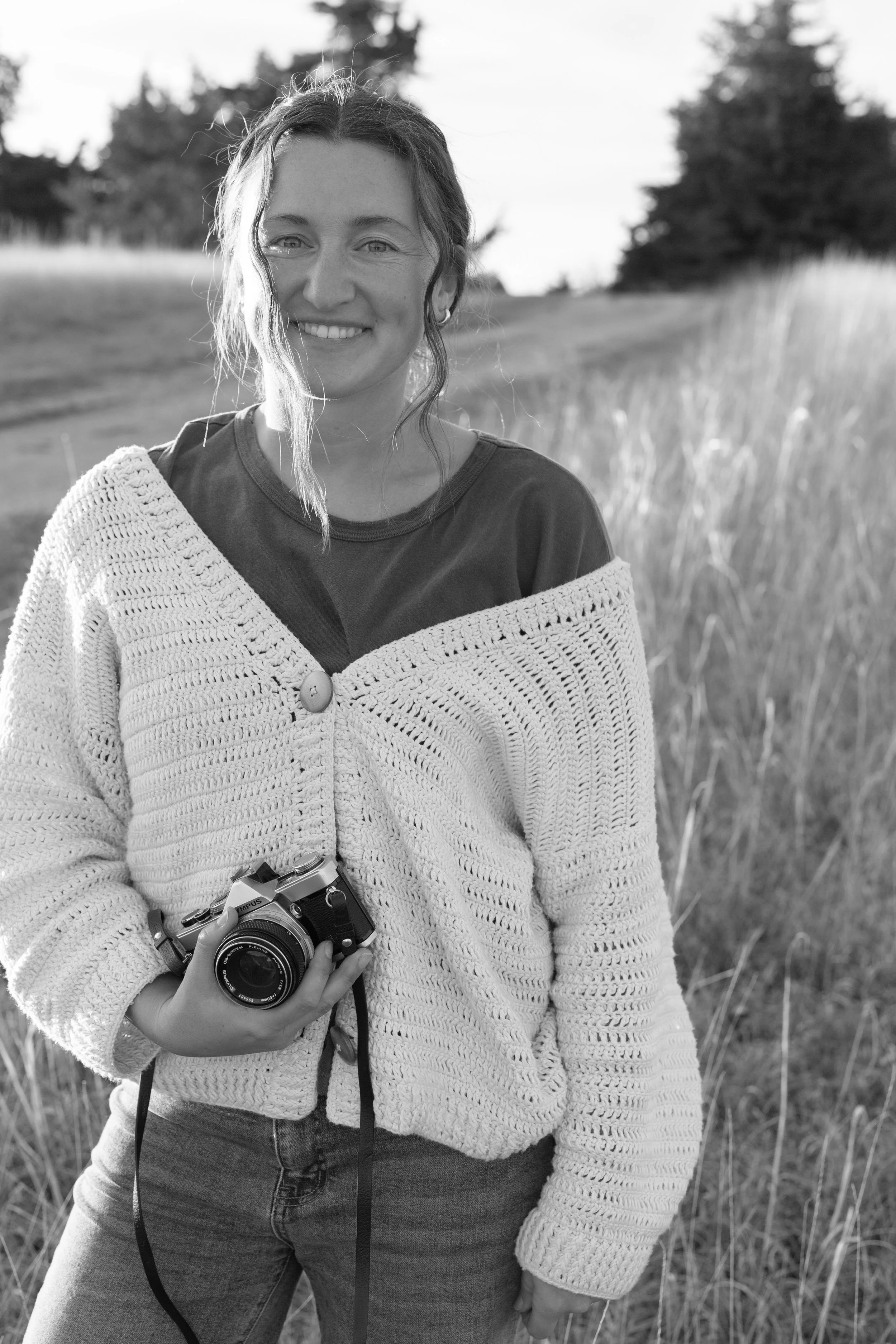 A woman standing outdoors in a field of tall, dry grass during sunset, holding a vintage-style camera, smiling at the camera.