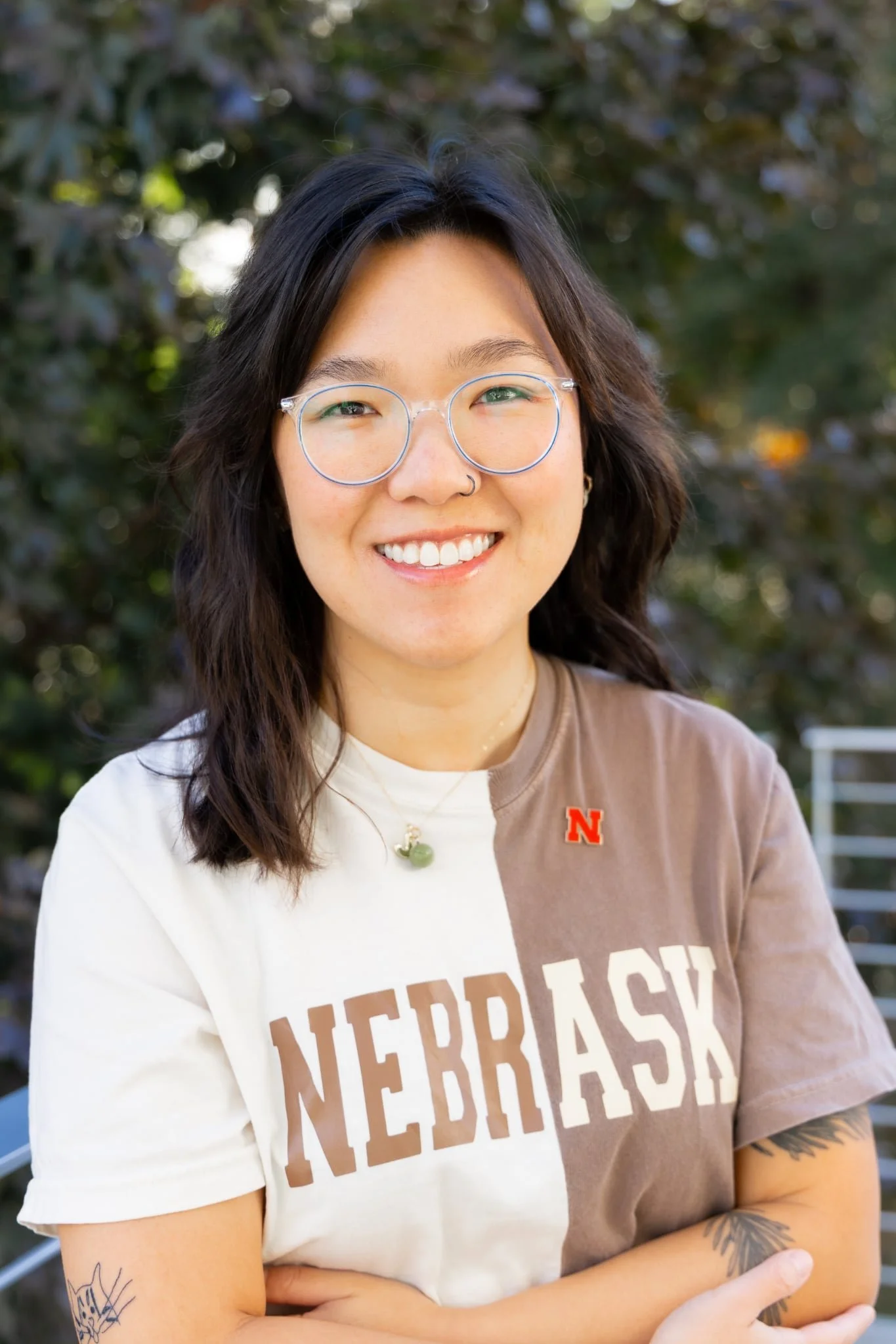 Smiling woman with glasses, nose ring, and tattoos wearing a Nebraska shirt outdoors.