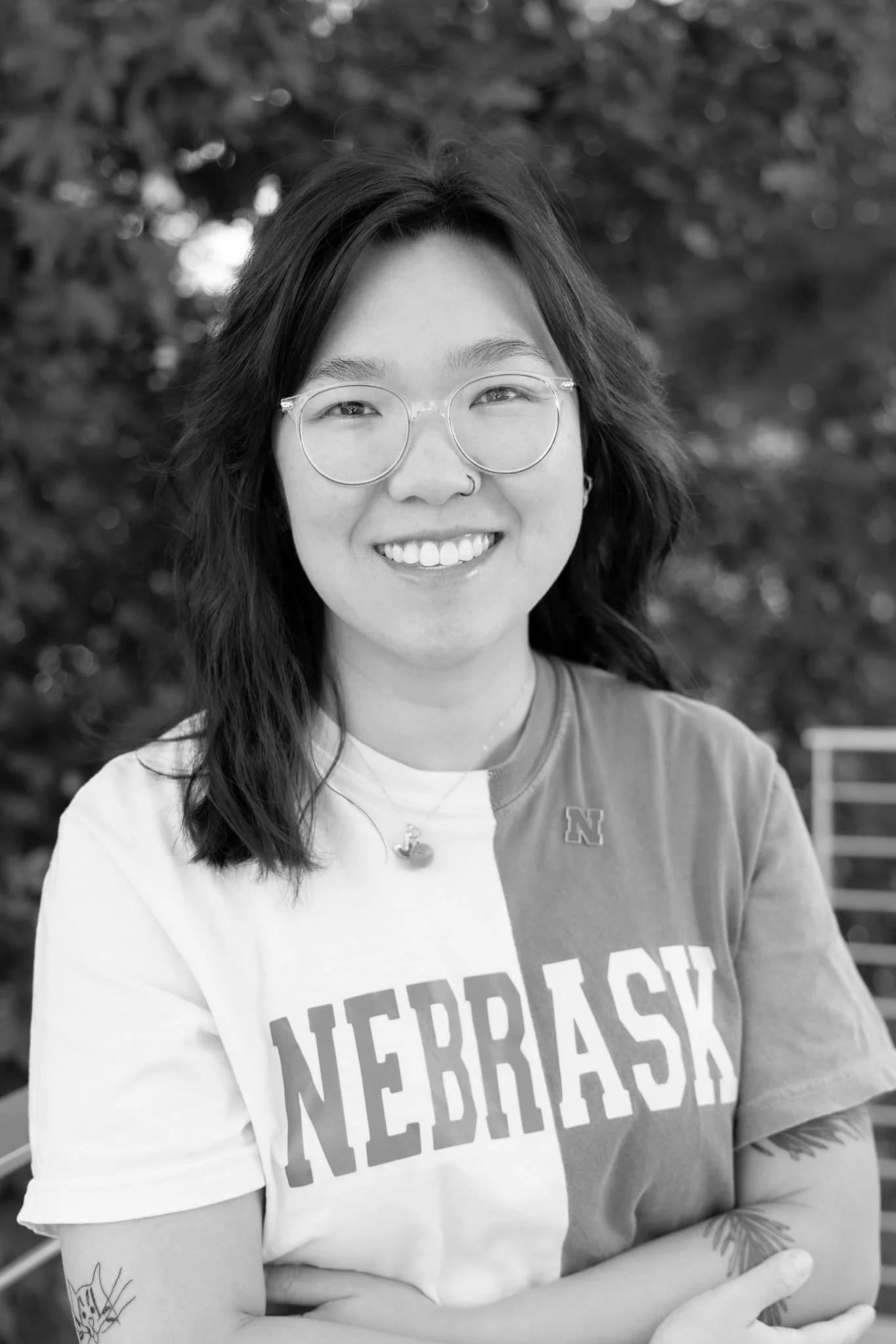 Smiling woman with glasses, nose ring, and tattoos wearing a Nebraska shirt outdoors.