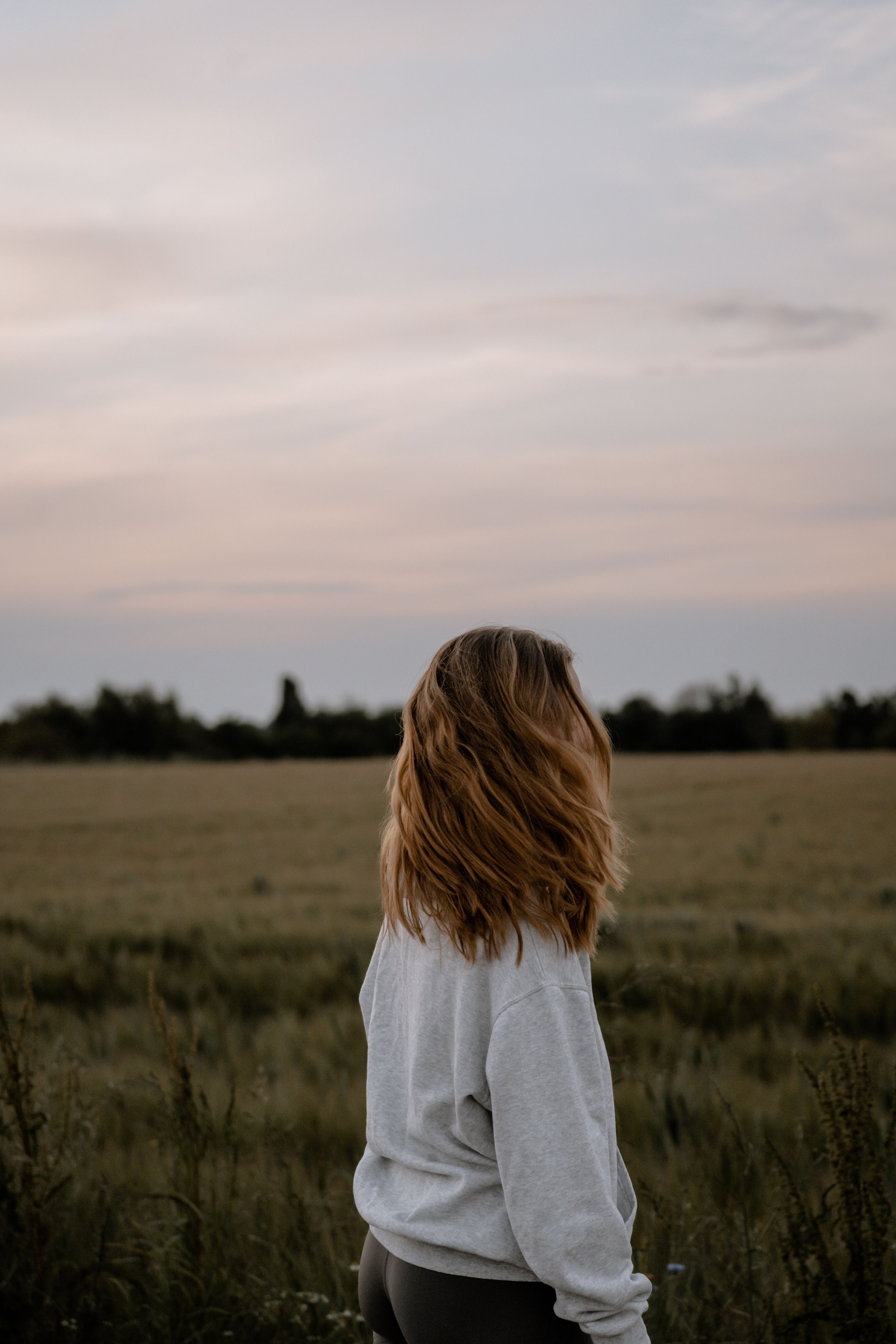 A girl with long, wavy auburn hair standing in a grassy field at dusk, wearing a gray sweatshirt and black leggings, facing away from the camera with a cloudy sky above.