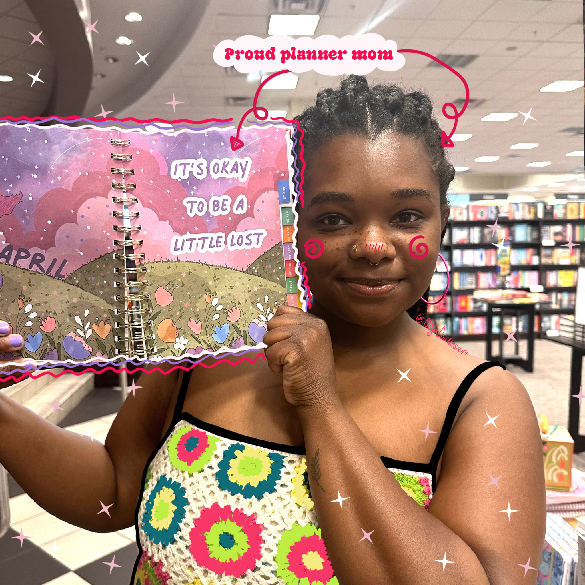 A woman with braids, wearing a floral dress, smiling in a bookstore, holding a pink and purple planner with the words 'It's okay to be a little lost' and 'Proud planner mom' with decorative sparkles and doodles added.