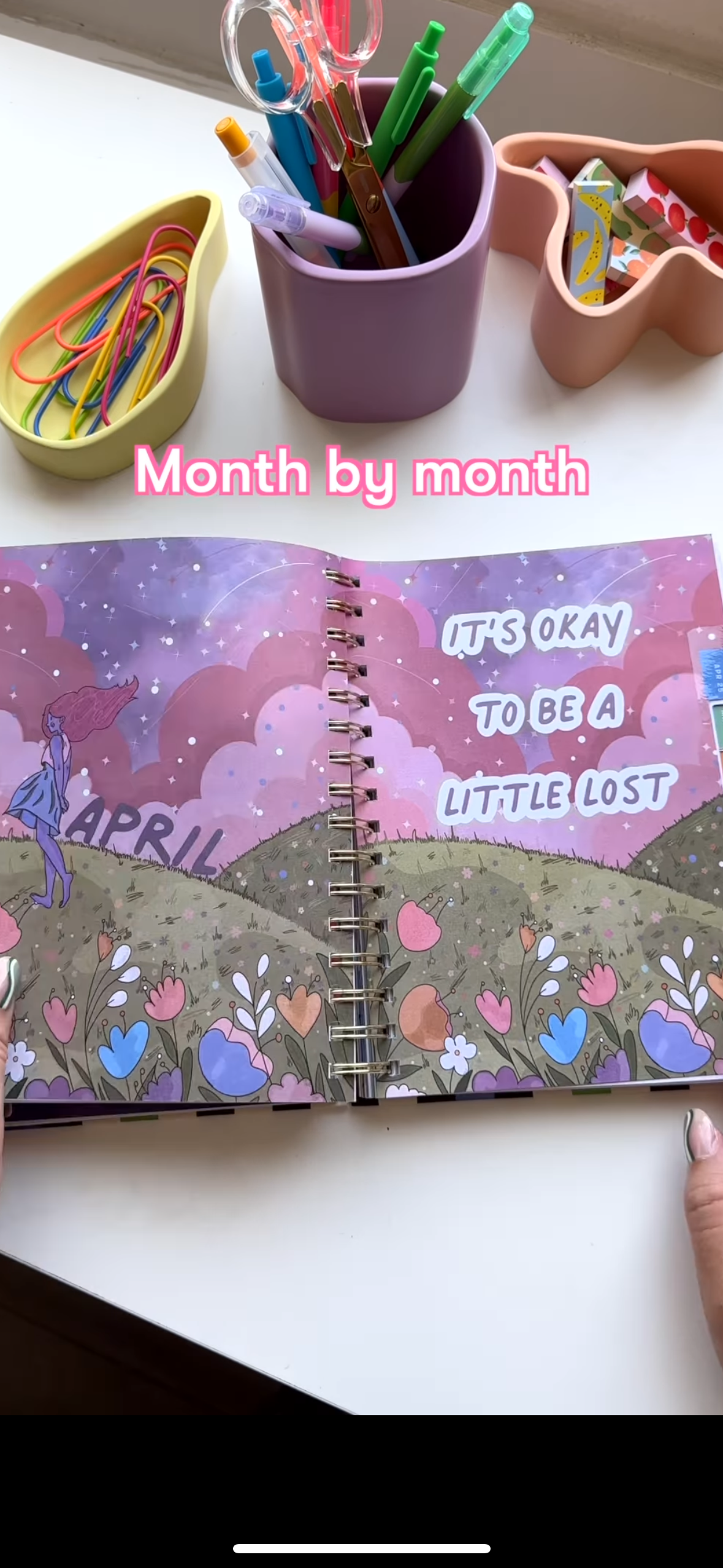 Open planner with illustrated month of April and motivational quote, surrounded by colorful stationery including paper clips, pens, transparent clips, and decorative tape on a white desk.