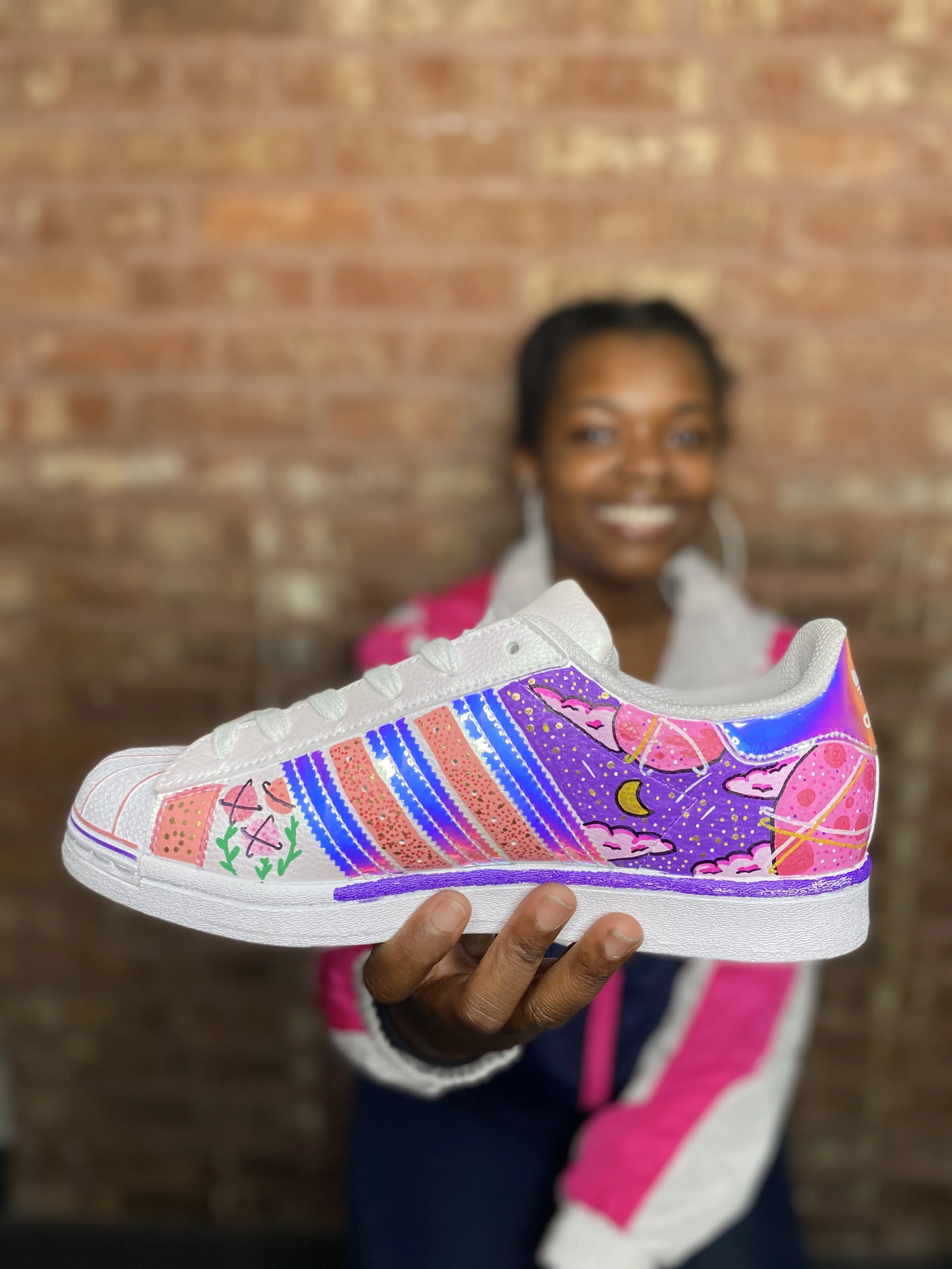 A smiling woman holding a white sneaker with colorful, artistic designs including pink, purple, blue, green, and yellow accents, featuring abstract and nature-inspired elements, against a brick wall background.