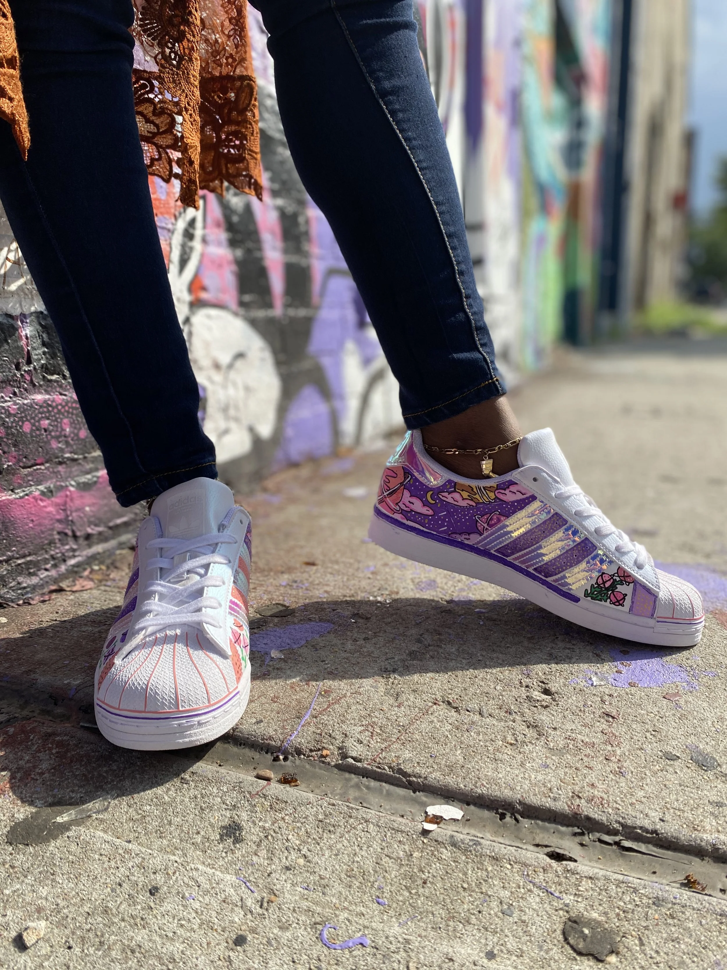 Person standing on sidewalk wearing white sneakers with colorful artistic designs and dark jeans, near a graffiti-covered wall.