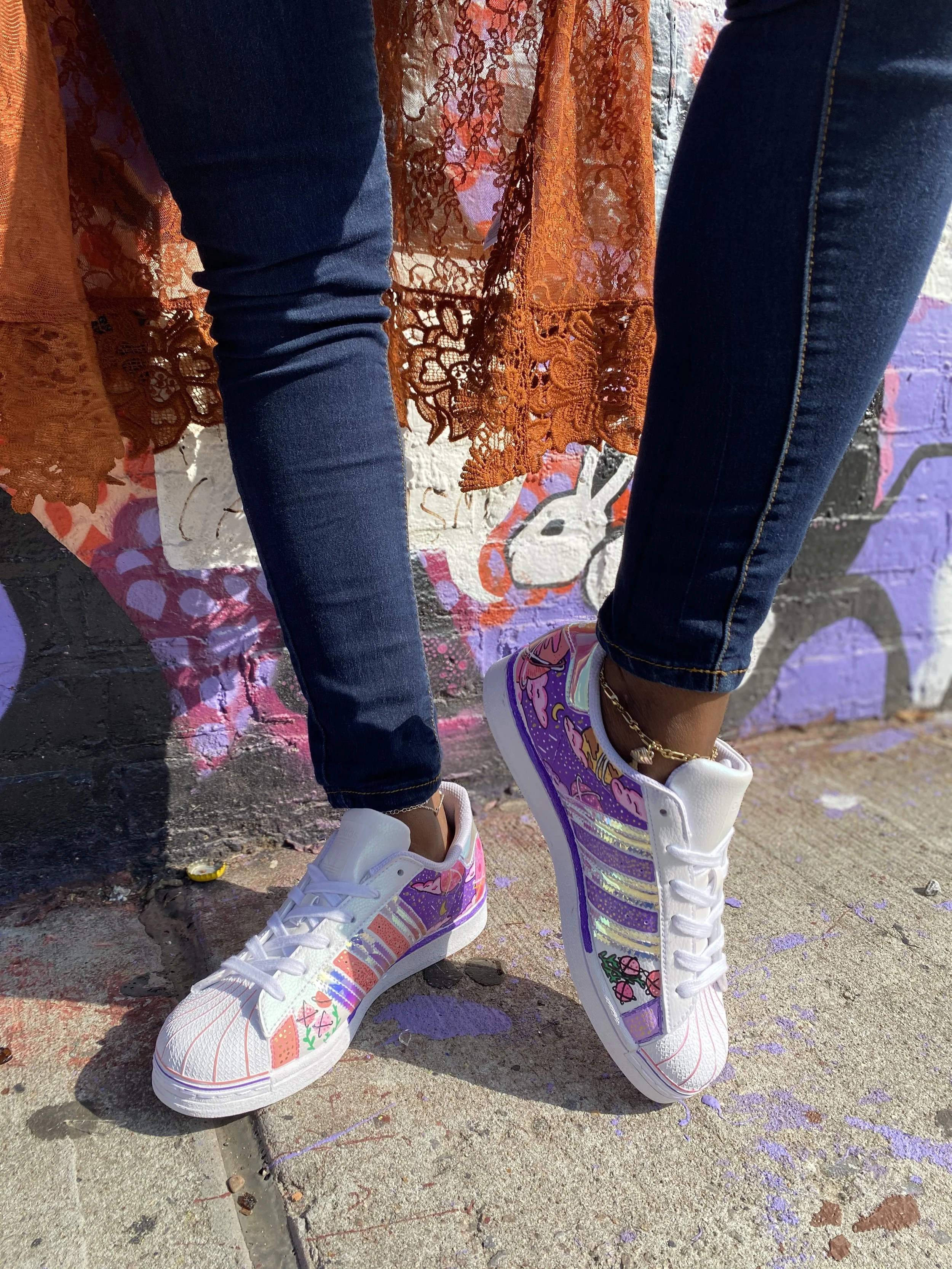 Close-up of a person wearing colorful, decorated sneakers with designs of cherries and strawberries, standing on a sidewalk near a graffiti-covered wall.