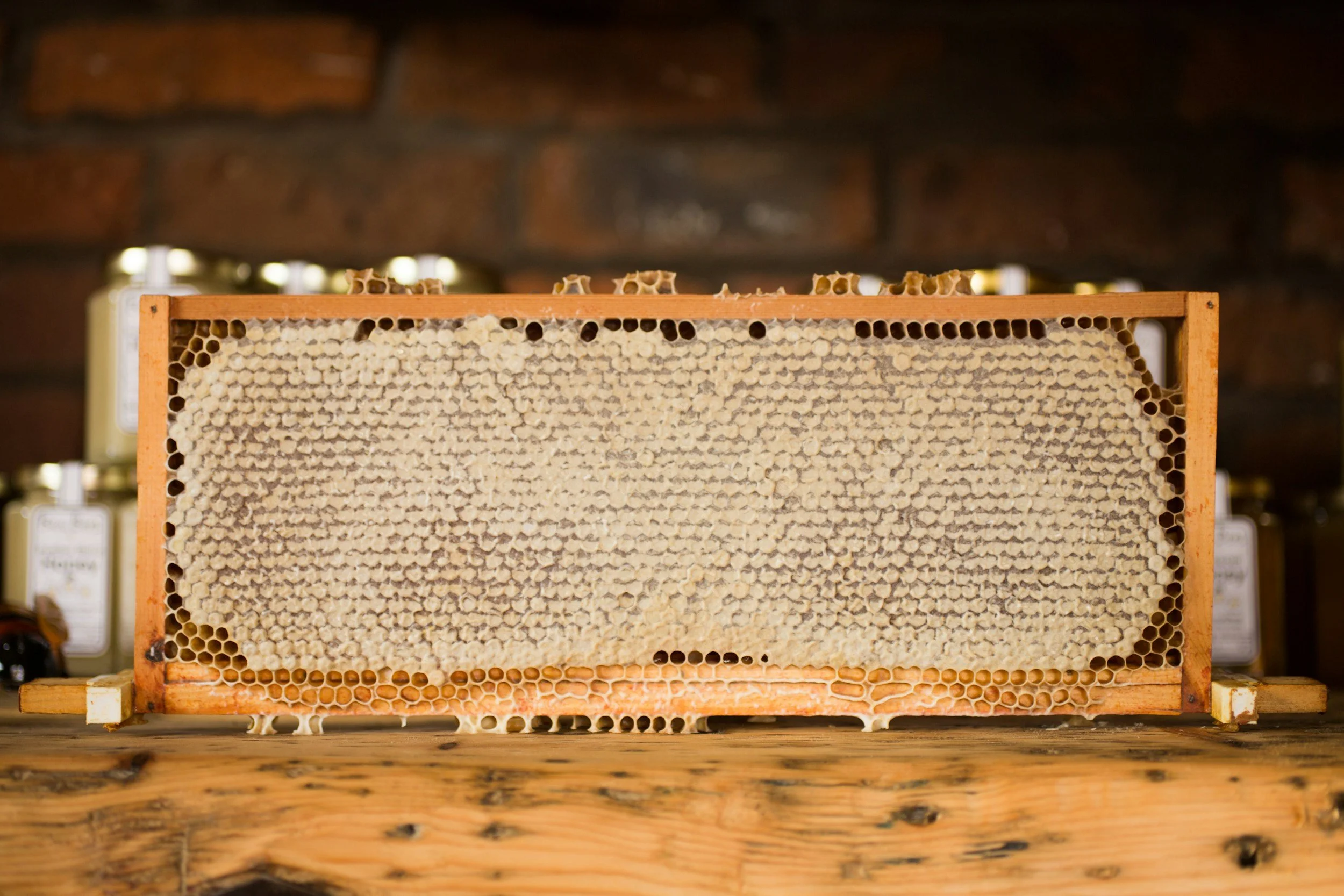 A honeycomb frame with beeswax cells, placed on a wooden surface with jars of honey in the background.