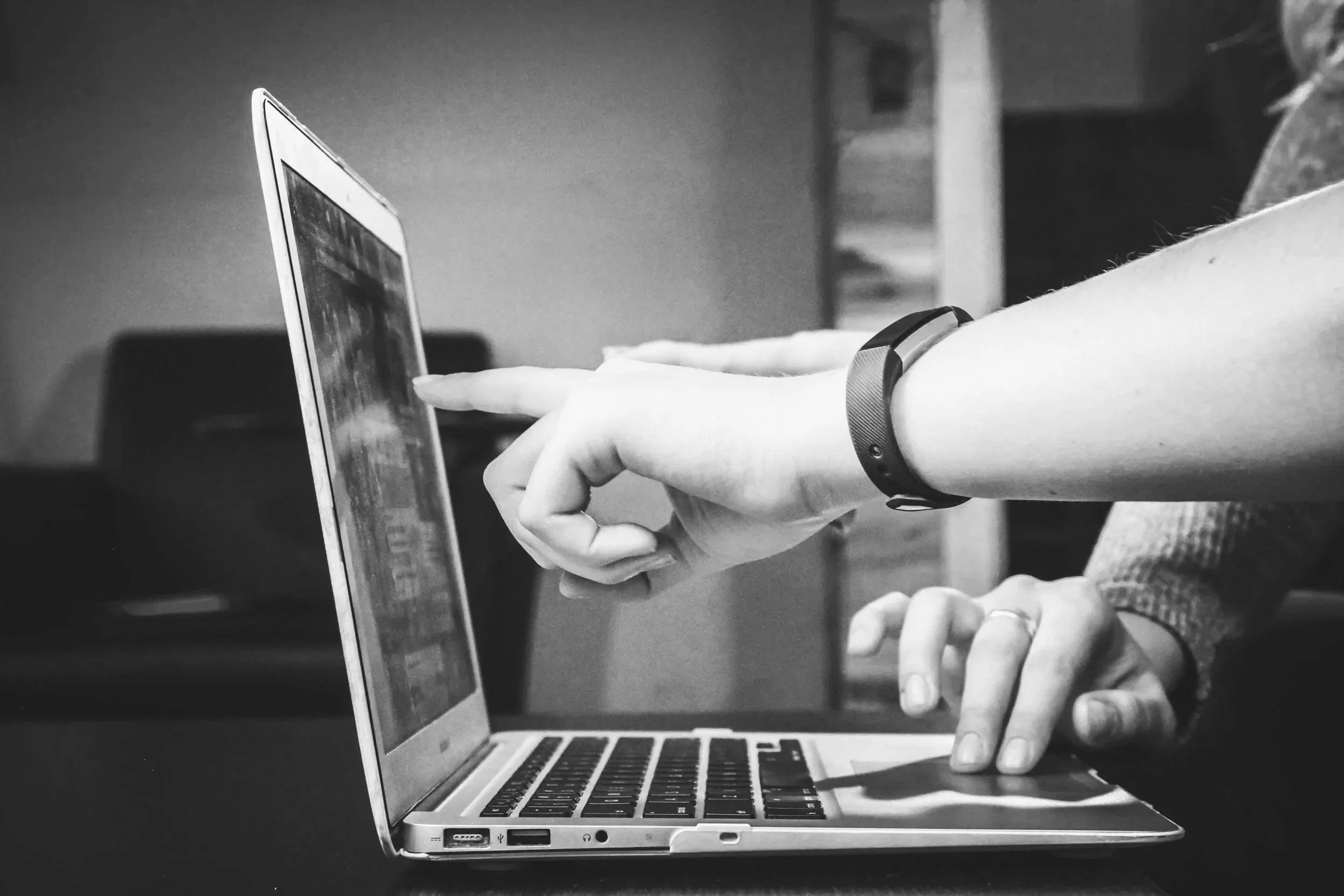 Black and white photo of a person's hand wearing a wristband, using their index finger to tap on a smartphone screen, while another hand supports the phone on a laptop keyboard.