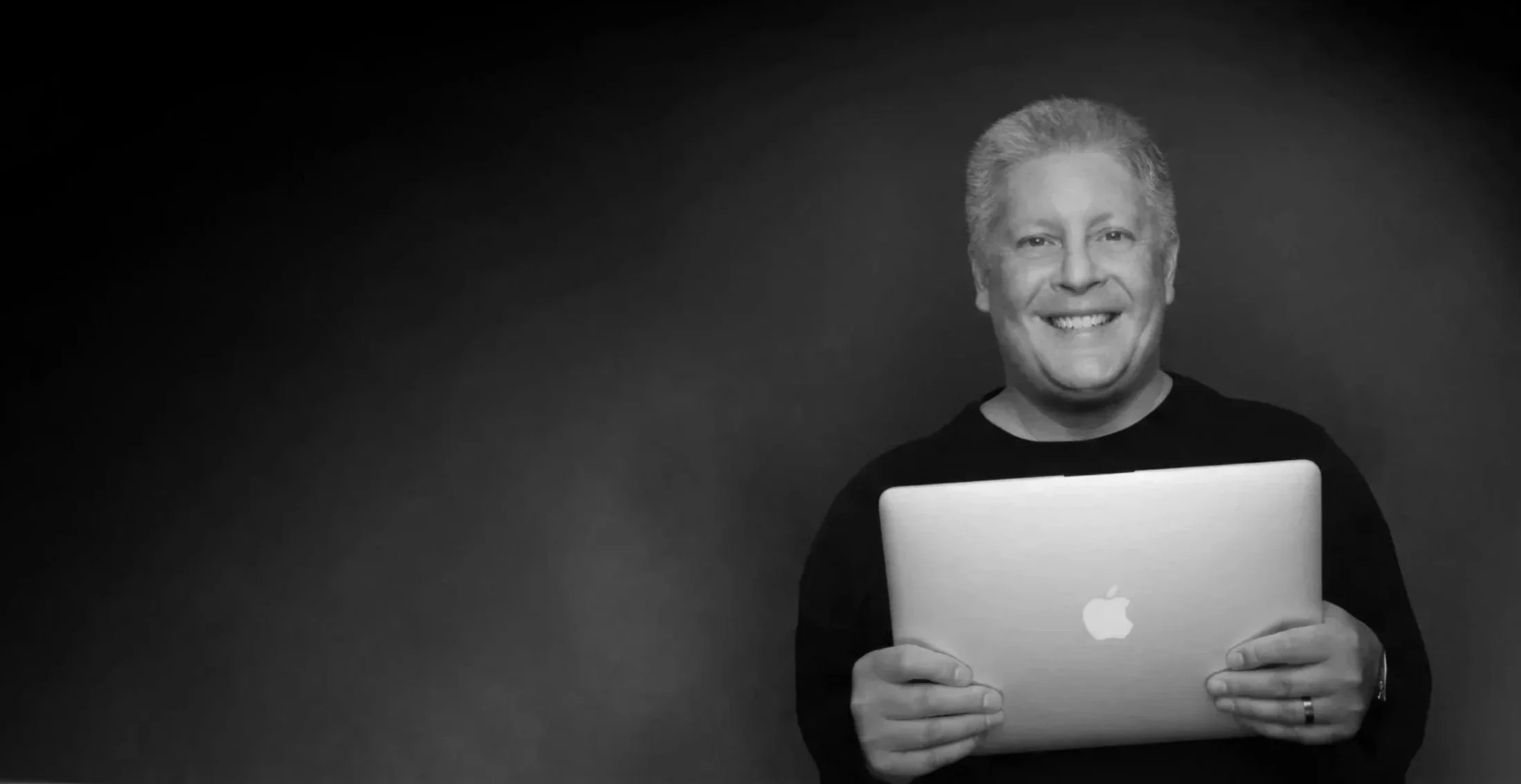 A smiling man with gray hair holding a silver Apple MacBook in front of a dark background.