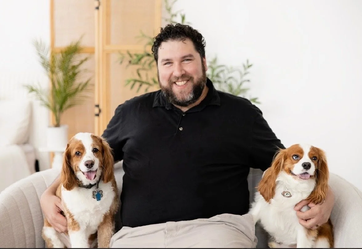 A man sitting on a couch with two dogs, smiling at the camera in a bright living room with plants in the background.