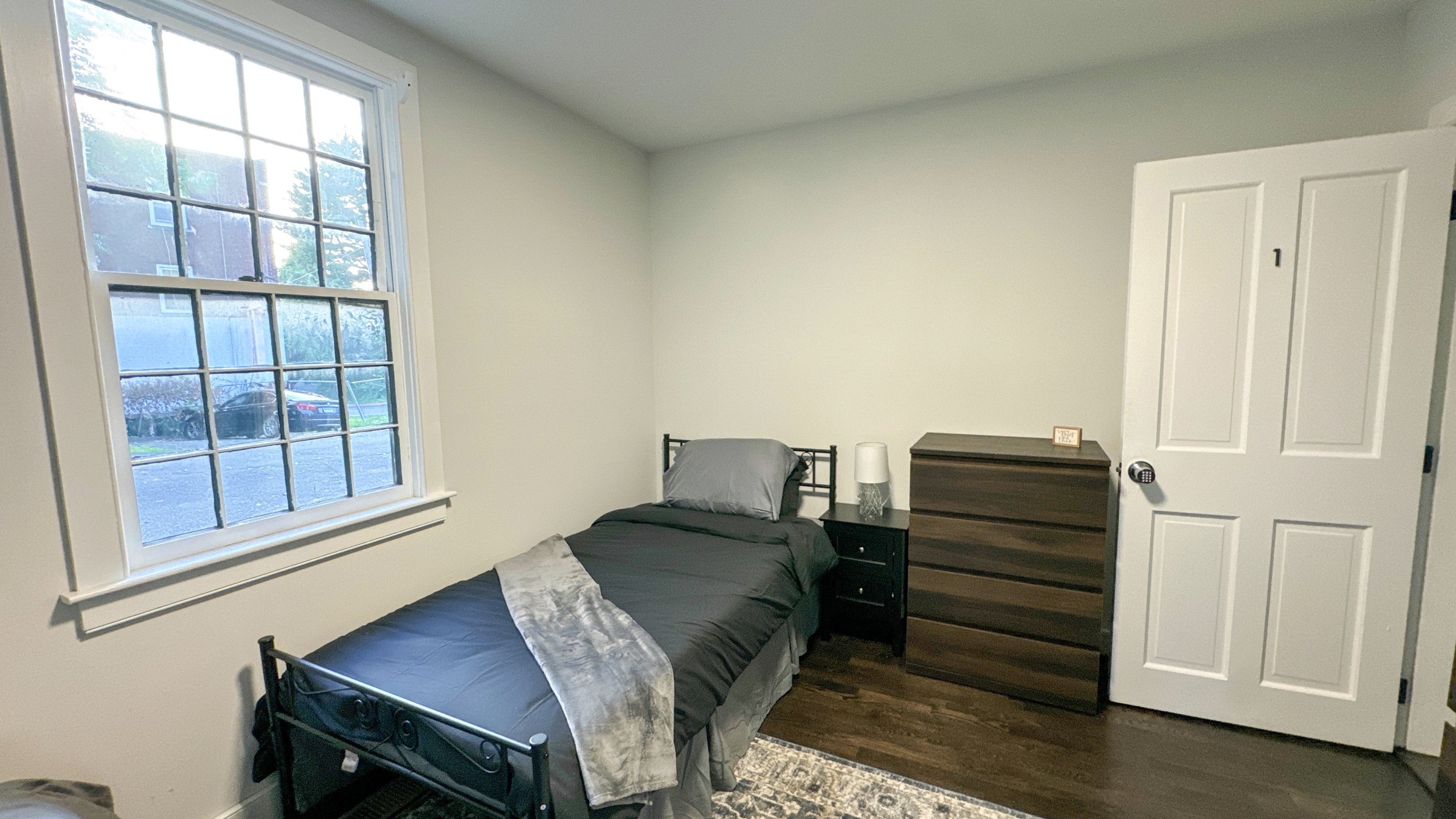 A small bedroom with a single bed, a black metal frame, gray bedding, a black nightstand with a white lamp, a brown dresser, a white door, and a window with grid panes showing an outside view of a car and trees.
