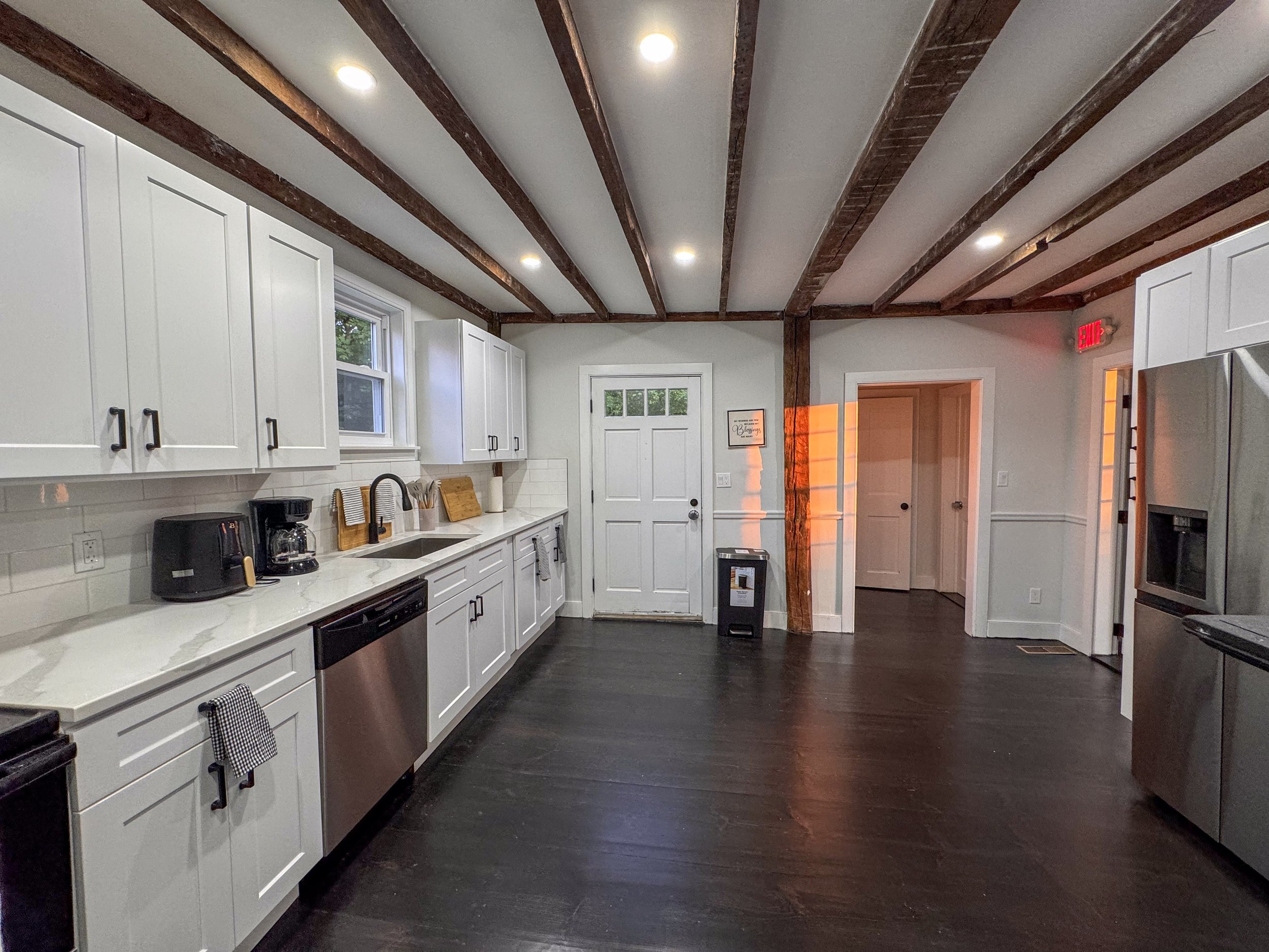 Modern kitchen with white cabinets, dark hardwood floors, and exposed wooden beams on the ceiling. Recessed lighting, stainless steel appliances, and a small window above the sink. There is a coffee maker, toaster, and set of cutting boards on the co