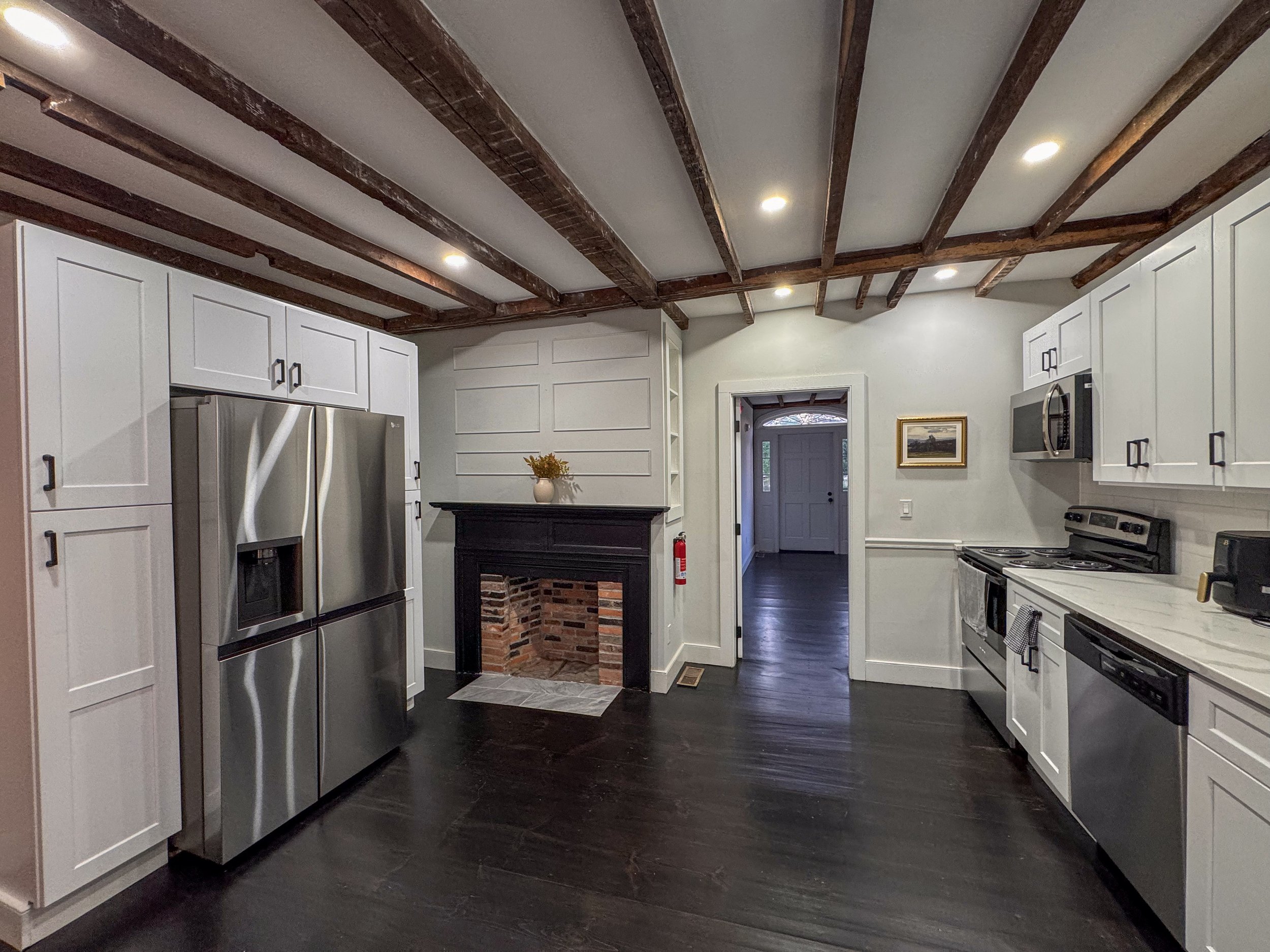Modern kitchen with white cabinets, stainless steel appliances, dark wooden flooring, exposed wooden ceiling beams, a small brick fireplace with a white mantel, and a hallway leading to a front door.