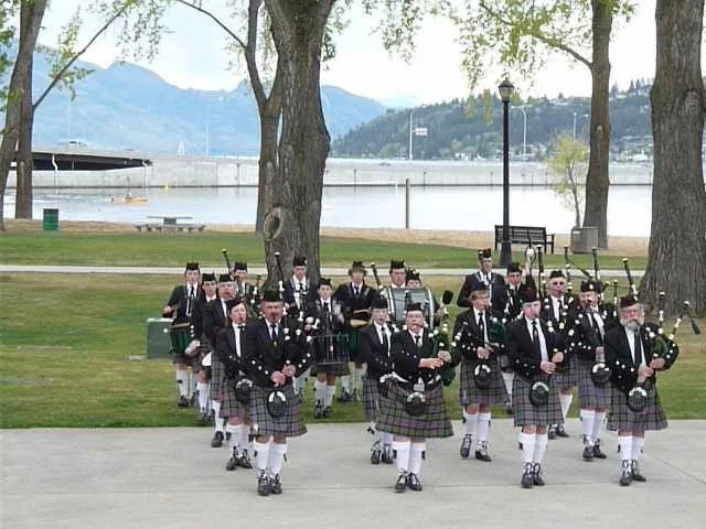 A marching band dressed in traditional Scottish attire, including kilts, performing outdoors near a lake with trees and mountains in the background.