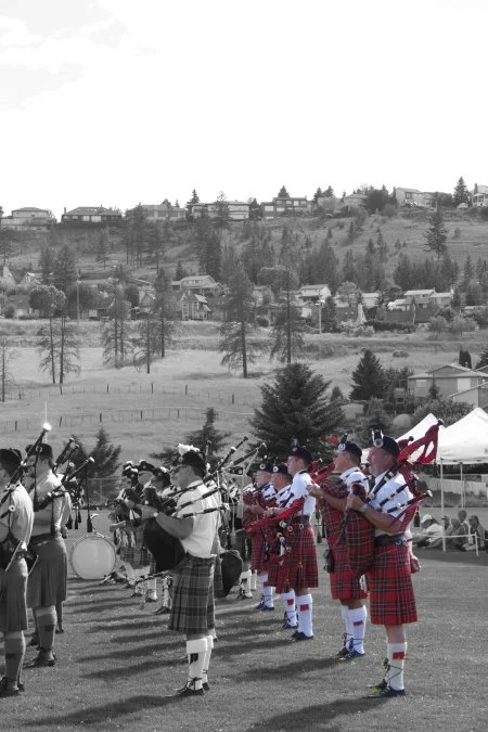 A group of people dressed in traditional Scottish attire, playing bagpipes and drums, performing outdoors on a grassy field during daytime, with a hillside and houses in the background.