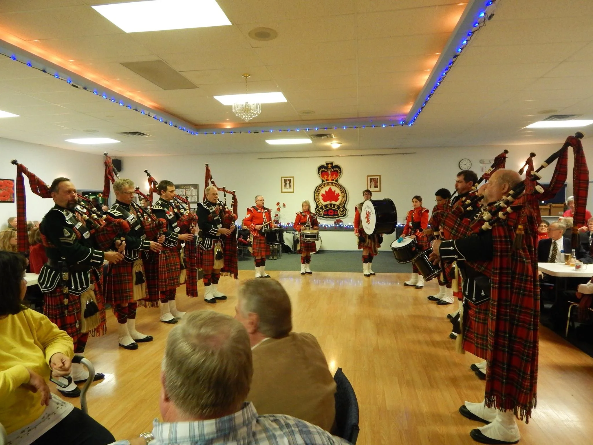 A group of musicians dressed in Scottish kilts and uniforms perform a pipe and drum concert in a banquet hall, with audience members watching.