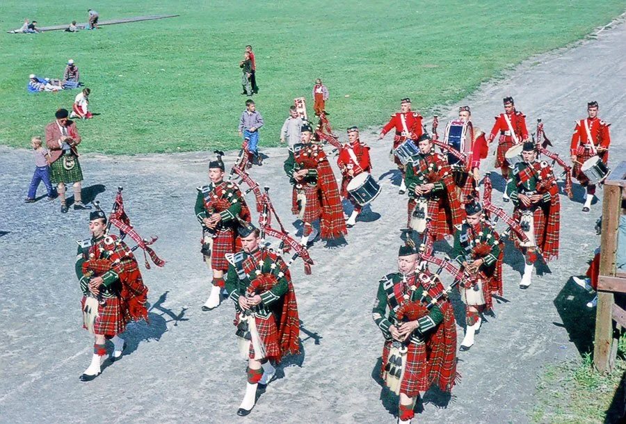 A group of people dressed in traditional Scottish attire, including kilts, playing bagpipes outdoors on a gravel path, with onlookers sitting on the grass in the background.