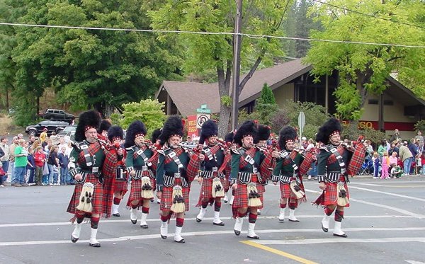 A marching band dressed in traditional Scottish attire, including kilts and bearskin hats, performing during a parade with spectators watching from the sidewalk.