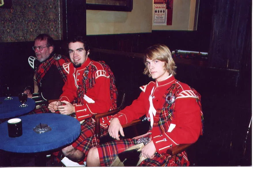 Three men sitting at a table dressed in traditional Scottish attire, including red jackets and tartan kilts, in a dimly lit pub.