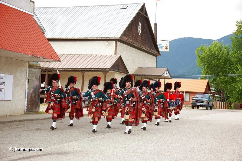 A marching band of musicians dressed in traditional Scottish attire, including kilts and bearskin hats, playing bagpipes and drums as they walk down a small town street.
