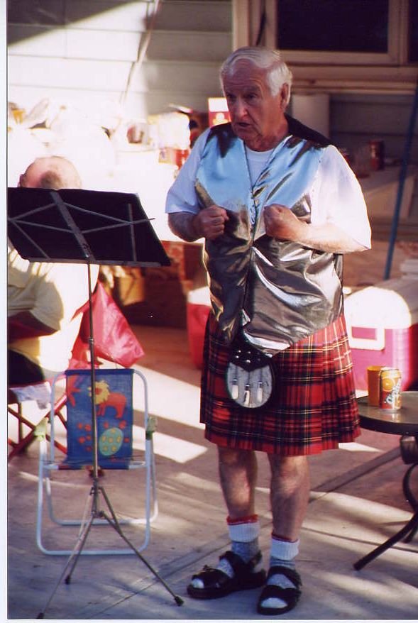 An elderly man with white hair wearing a shiny silver vest, plaid shorts, and sandals with socks, standing outdoors near a music stand, possibly at a gathering or event.