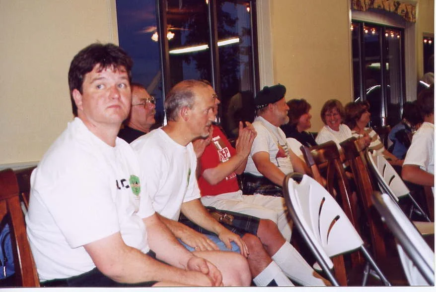 A group of people sitting on chairs in a room, some looking towards the front and others looking away, with windows showing the evening outside.