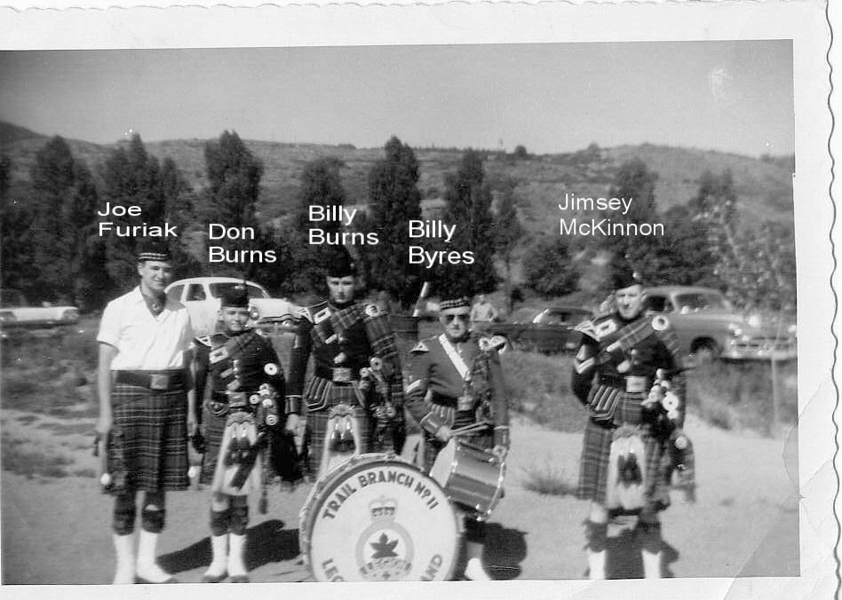 Historical black and white photo of five men in Scottish Highland dress standing outdoors with cars and a hilly landscape in the background. Four of the men hold bagpipes, and one has a drum with the text "Trail Braml M111".