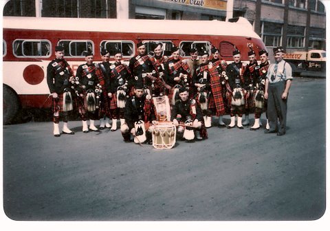 Group of men in kilts and uniforms standing and kneeling in front of a vintage bus, with a few small drums on the ground, in an urban street setting.