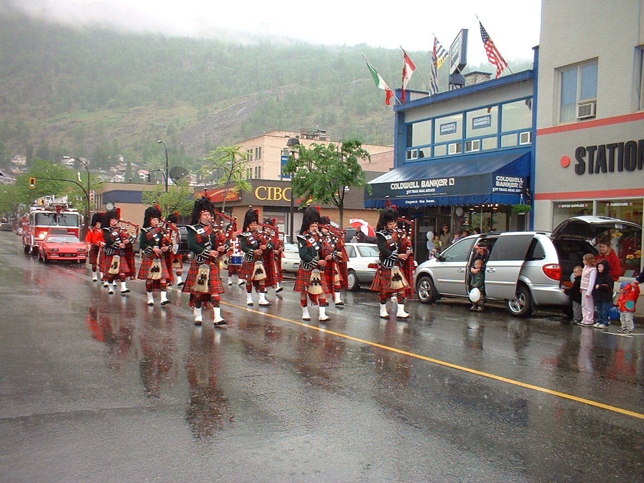 A parade featuring a marching band dressed in traditional Scottish attire, playing bagpipes and drums, on a rainy street with onlookers and parked cars.