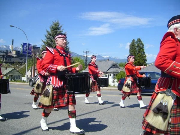 A group of people dressed in traditional Scottish attire, including red jackets and tartan kilts, marching in a parade and playing drums on a street.