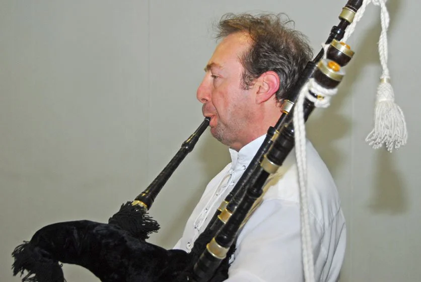 A man playing a bassoon, viewed from the side, with a gray background.