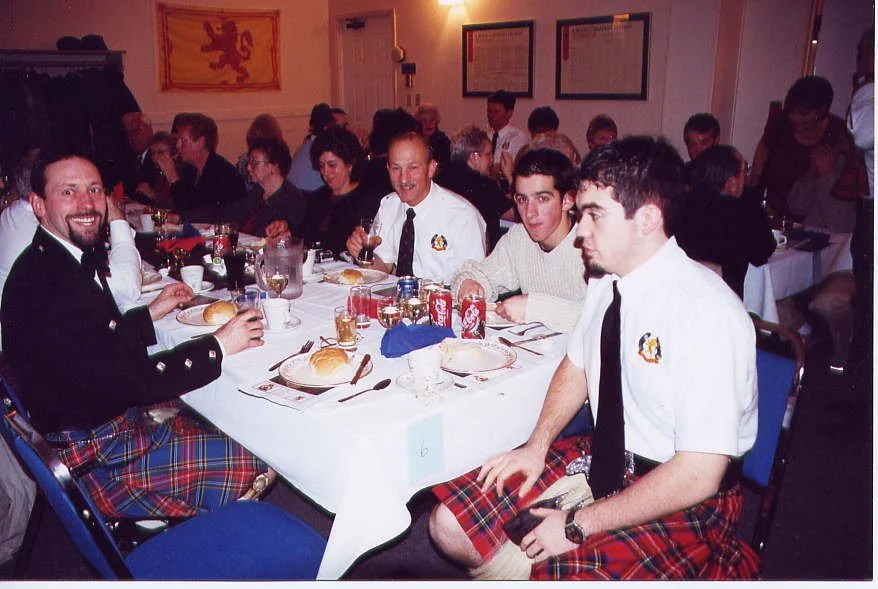 Group of men dressed in traditional Scottish attire, including kilts, enjoying a meal and drinks at a banquet table.
